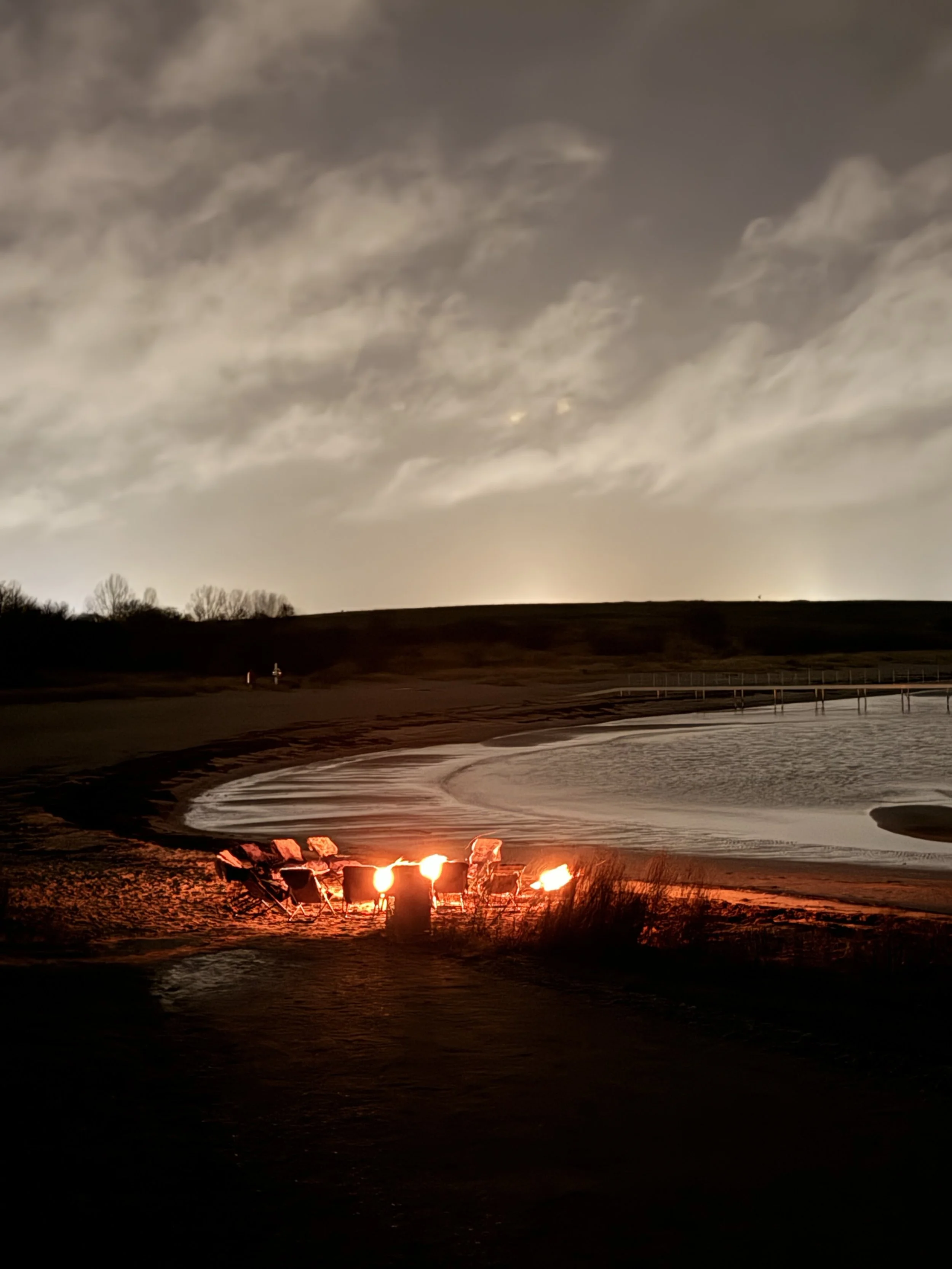 A beach at night with a fire and chairs arranged near the water, cloudy sky overhead.