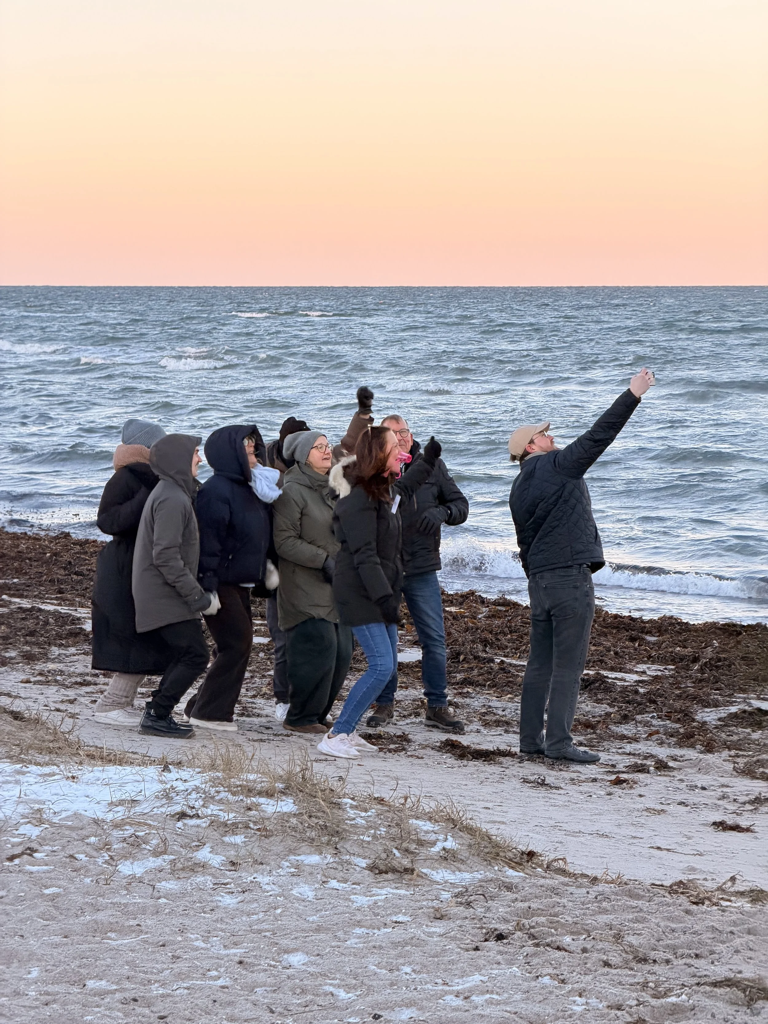 En gruppe mennesker tager selfies på stranden med havet i baggrunden ved solnedgang.