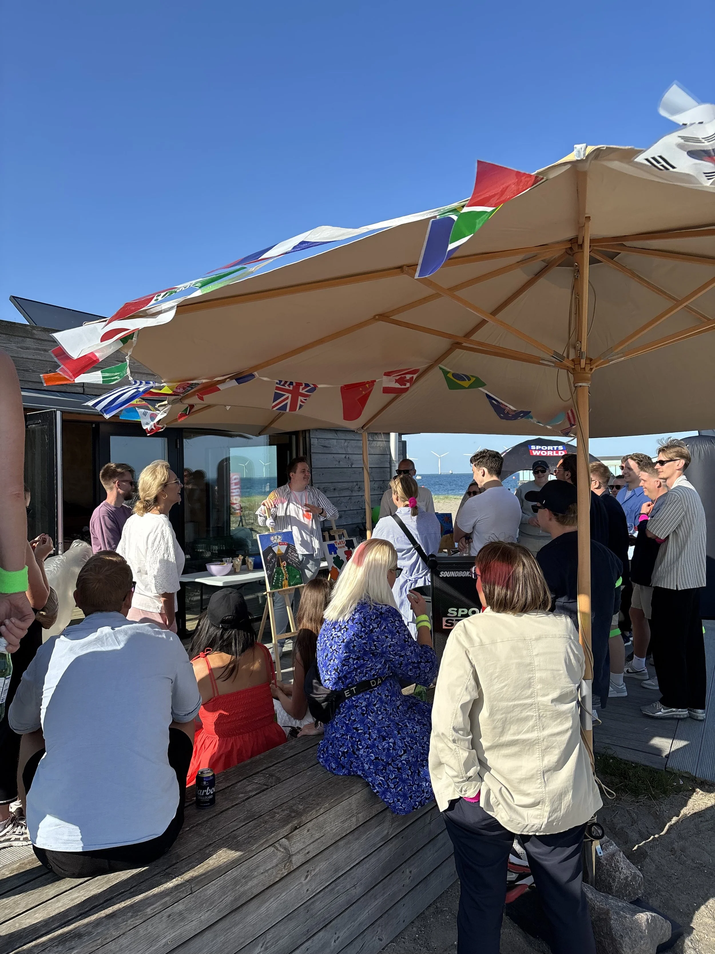 Udendørs sommerarrangement ved stranden i København med Sports World-bannere, parasoller og glade mennesker. Solrig dag med blå himmel, balloner og social stemning foran træbygninger i Amager Strandpark.