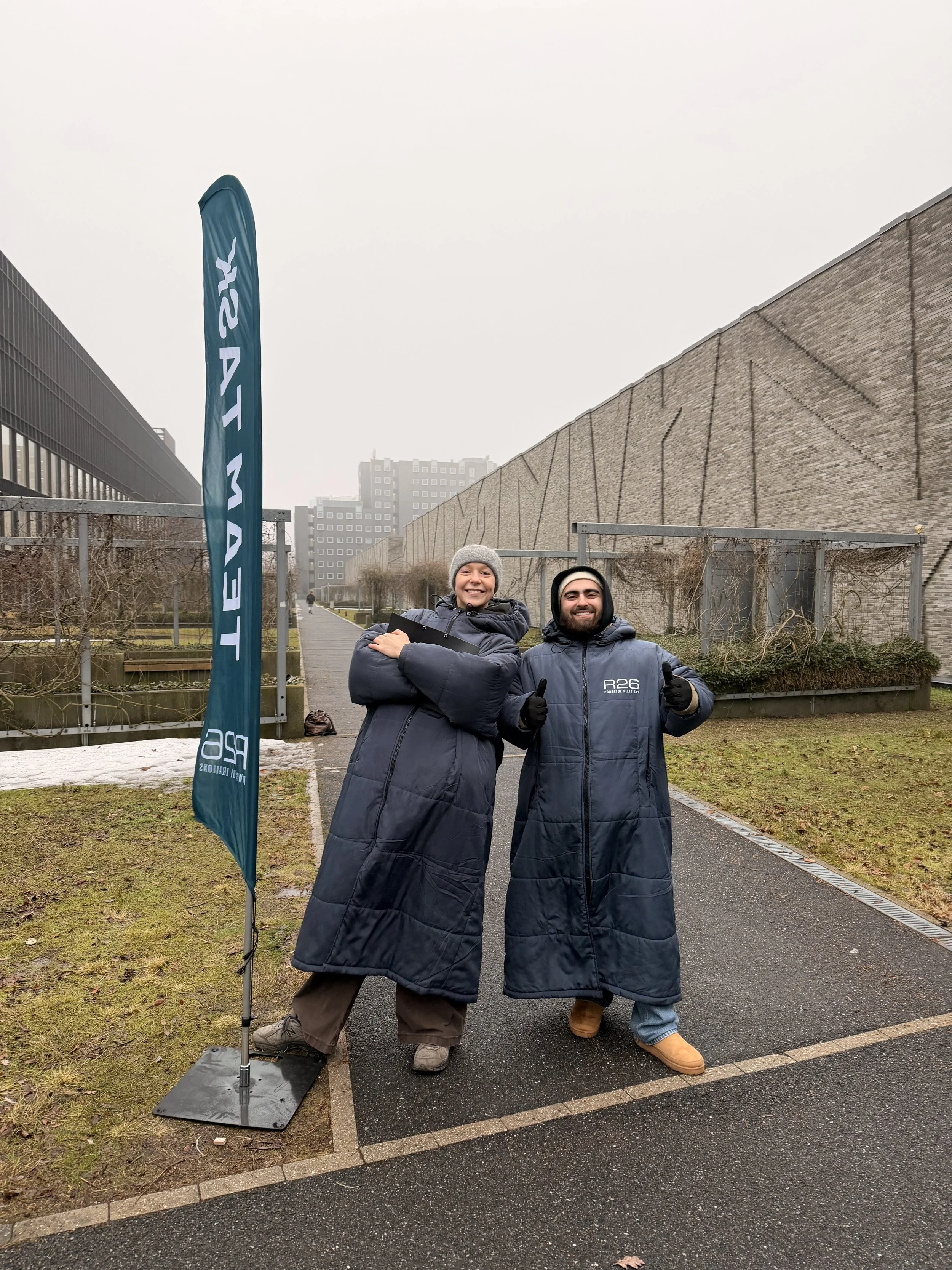 To personer står ude i regnen udenfor, begge iført lange, blå nattrokkener, og smiler til kameraet, med en blå flag med teksten 'RE 26' ved siden af.