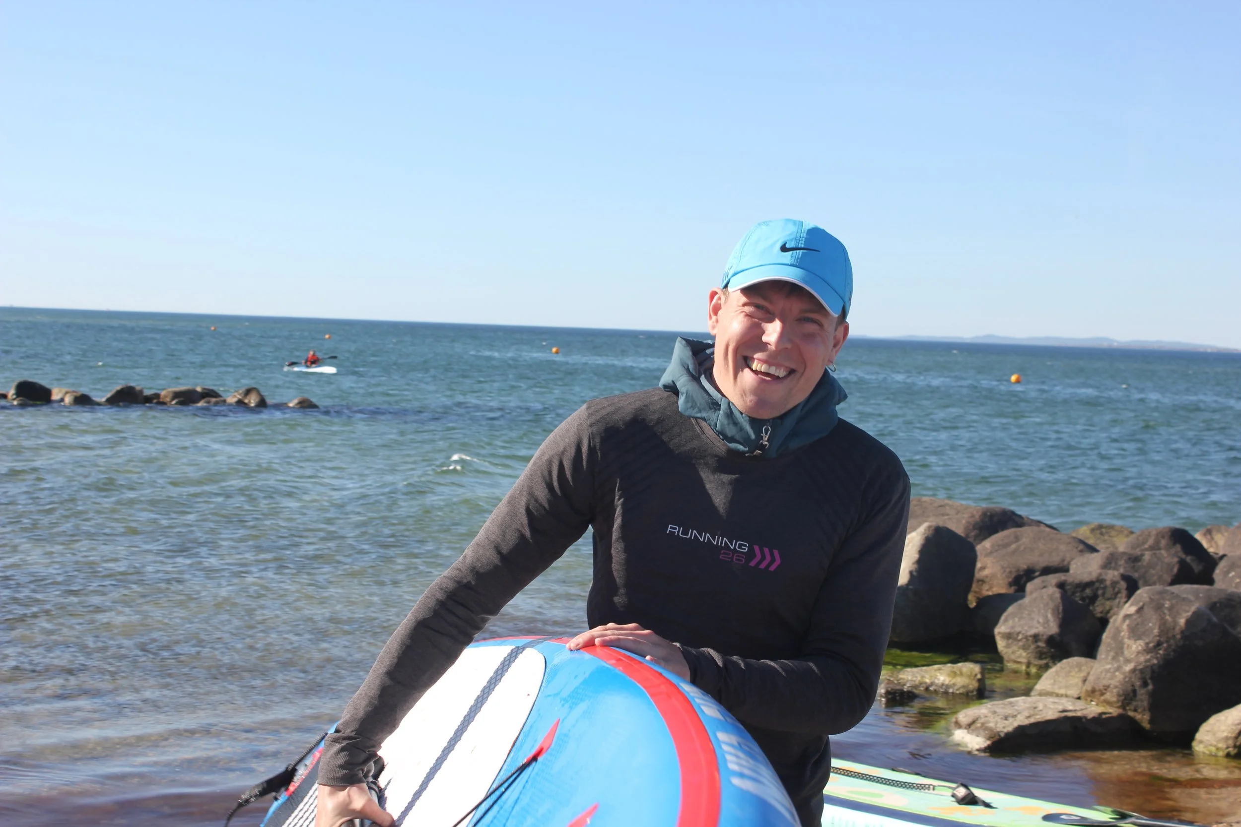 Smilende mand med blå cap og sort løbetrøje holder et blå-hvid-orange surfbræt ved kysten med sten og hav i baggrunden.