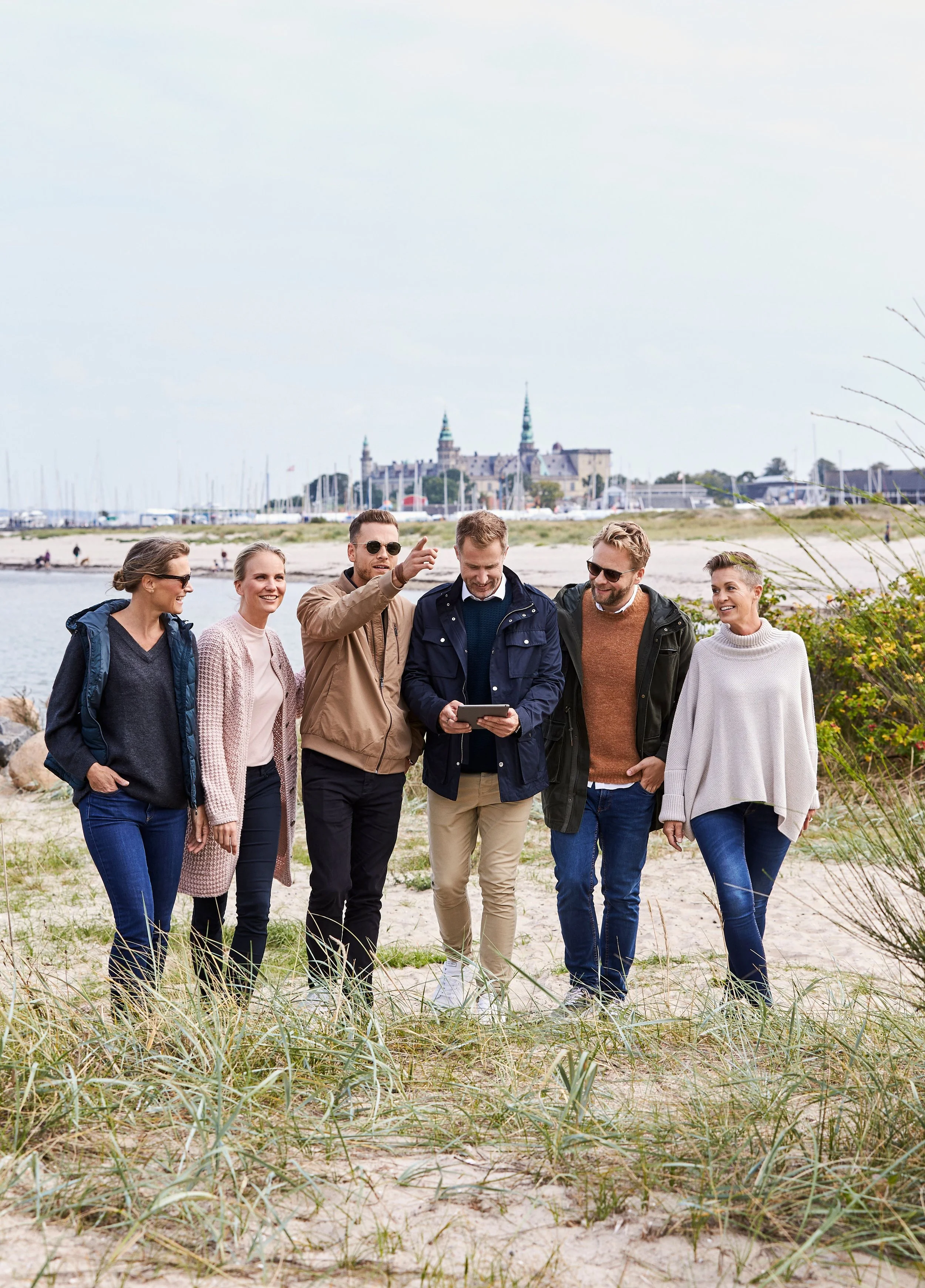 Gruppe af seks venner går på stranden og ser på en mobiltelefon, med en marina og slot i baggrunden.