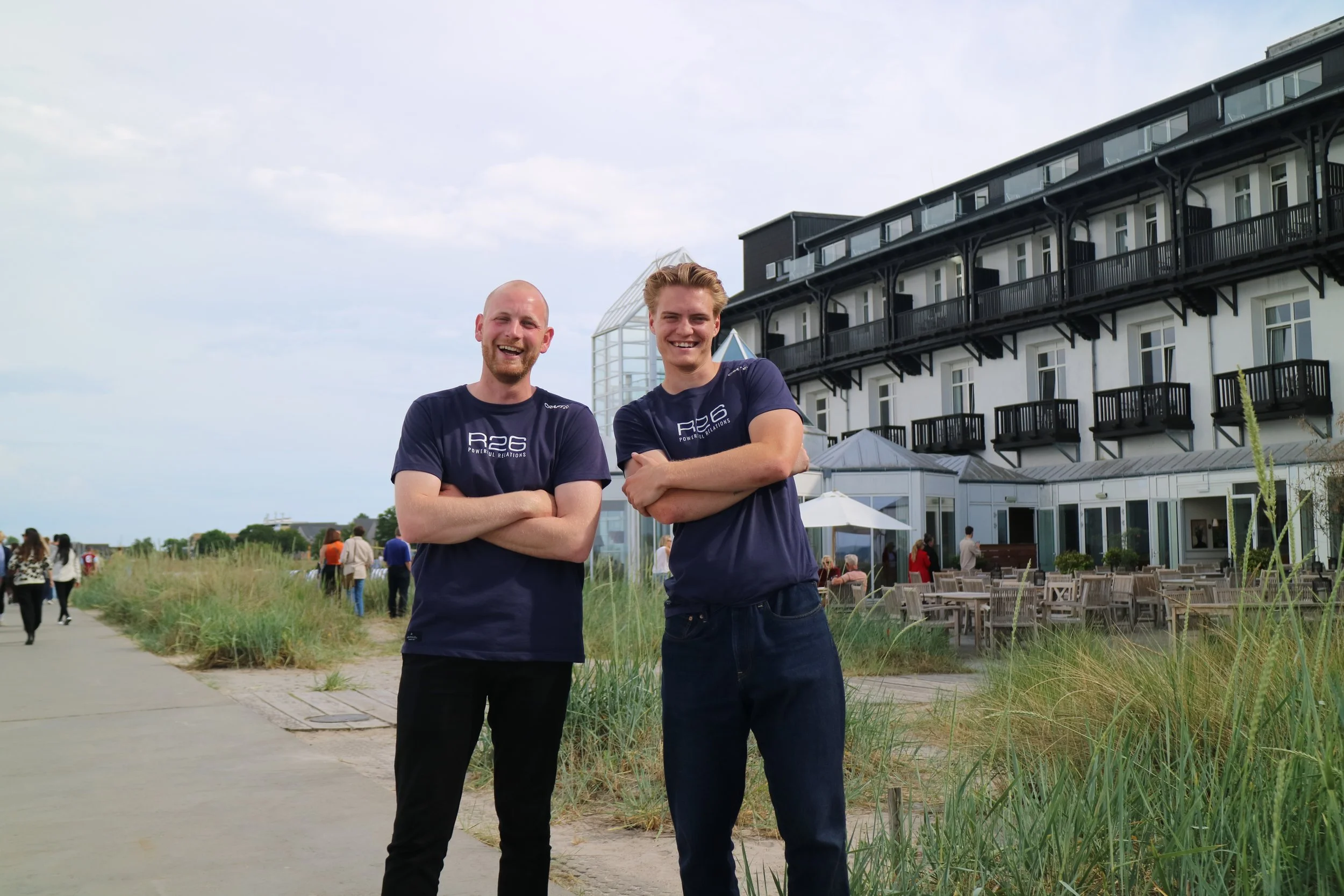 To unge mænd smiler og står med arme kryds foran en bygning ved stranden. Der er mennesker i baggrunden, og der er udendørs siddepladser og græsarealer.