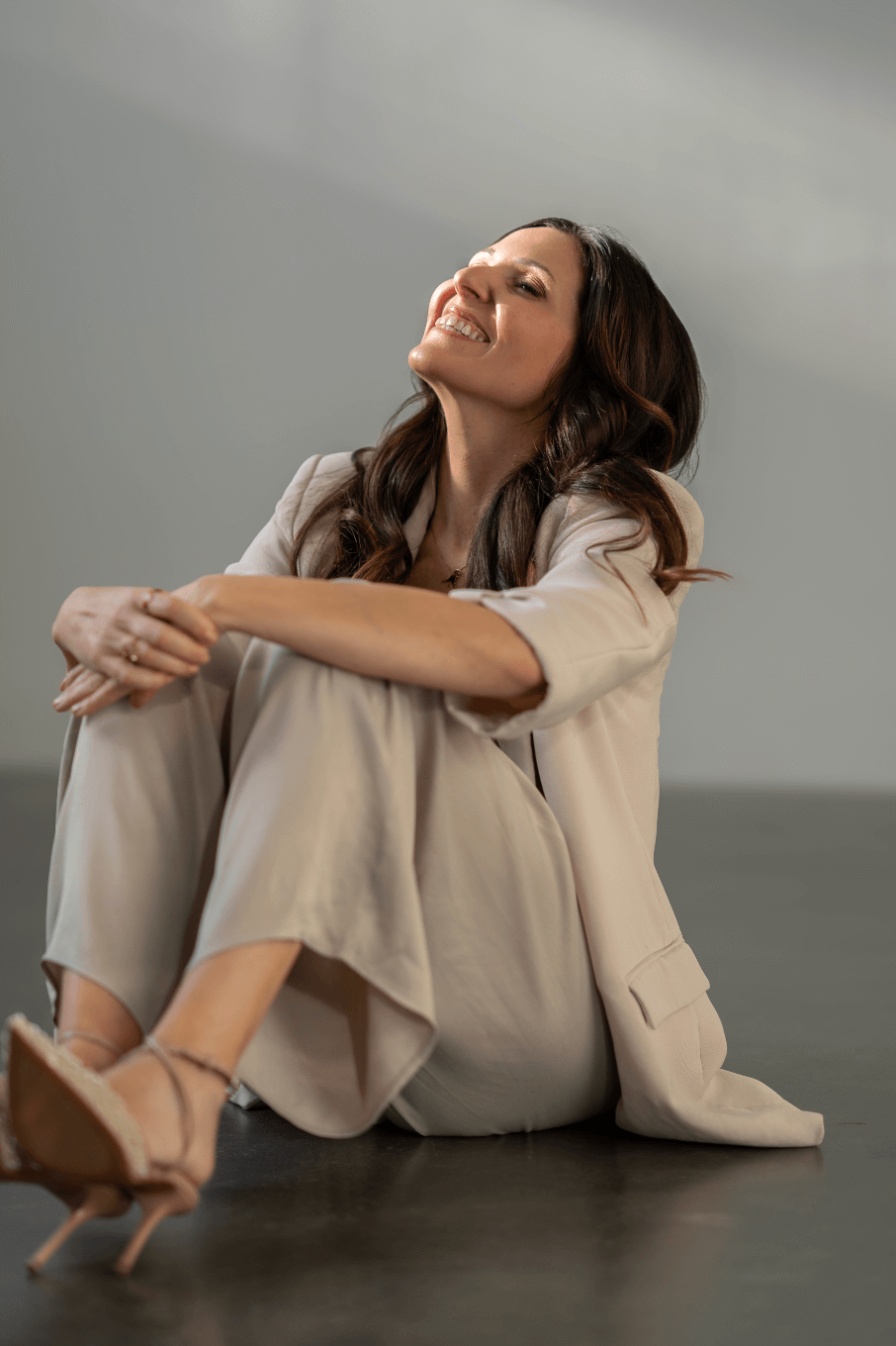 Eine lächelnde Frau sitzt auf dem Boden, trägt einen beigefarbenen Anzug und spitze Schuhe, und blickt glücklich nach oben.