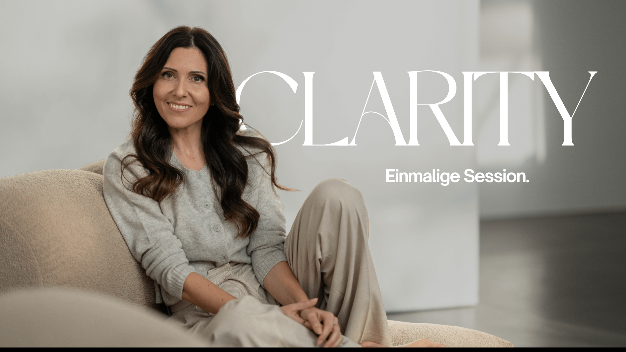 Eine lächelnde Frau mit langen, braunen, gewellten Haaren sitzt auf einer beige Couch in einem modernen, hellen Raum, getragen in einem grauen Cardigan und bequemen Hosen. Über der Frau steht in großen weißen Buchstaben 'CLEARTY' und darunter in kleineren Buchstaben 'Einmalige Session.'
