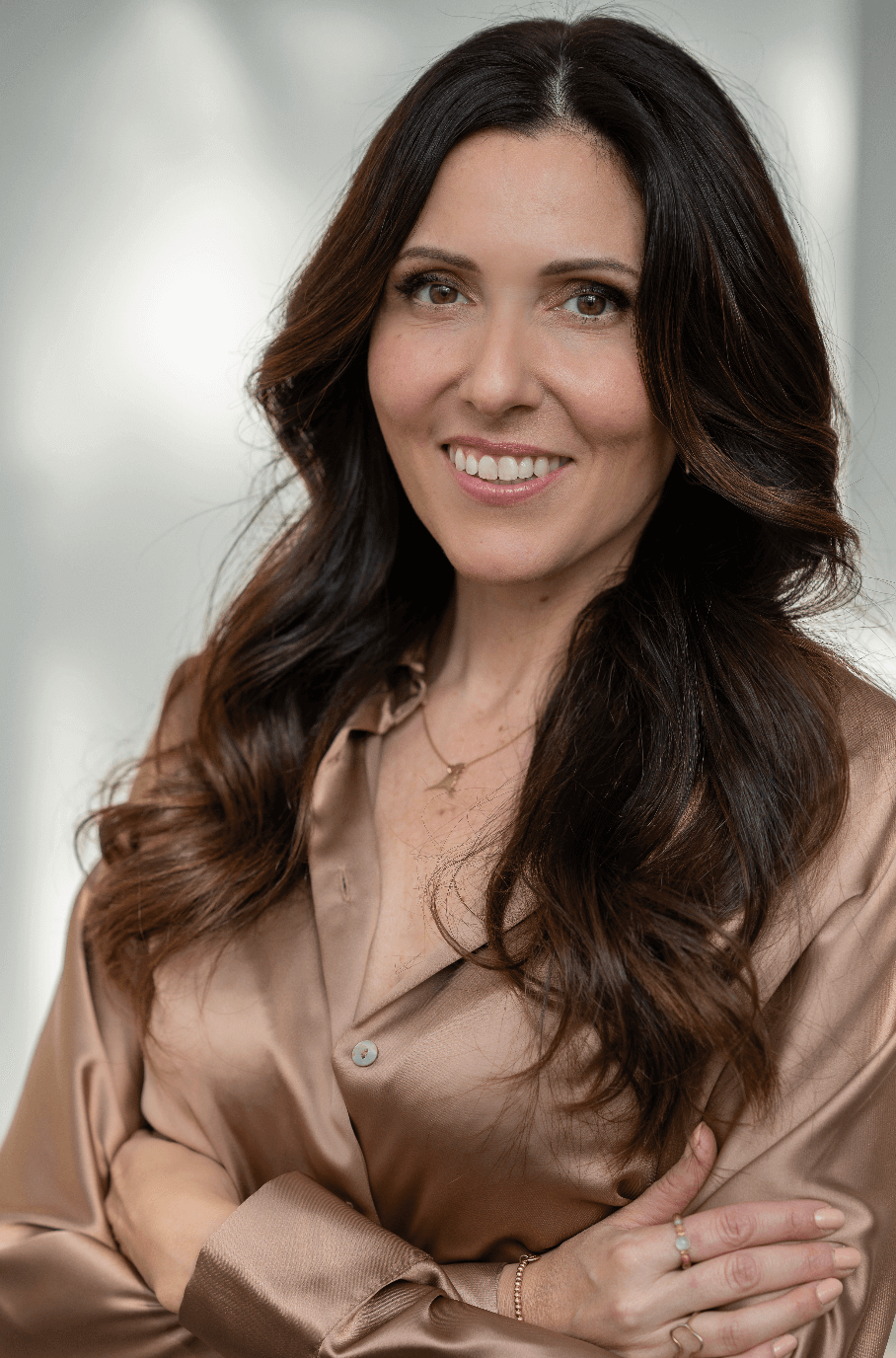 Eine lächelnde Frau mit langen, braunen, gewellten Haaren trägt eine beige Seidenbluse und eine zarte Halskette, steht vor einem neutralen Hintergrund.