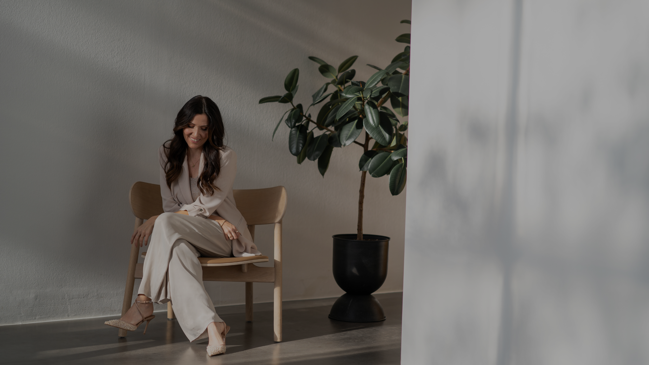 Eine Frau sitzt auf einem Holzsitz, lächelt und schaut nach unten, neben ihr steht ein großer grüner Pflanzenstrauch in einem schwarzen Topf, im Hintergrund eine weiße Wand.