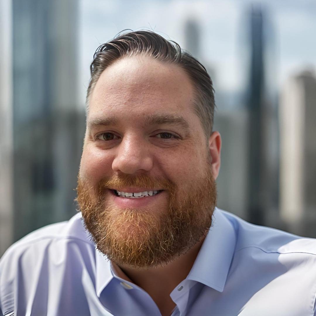 A smiling man with a beard and short, slicked-back hair wearing a light blue dress shirt, in front of a cityscape with tall buildings.