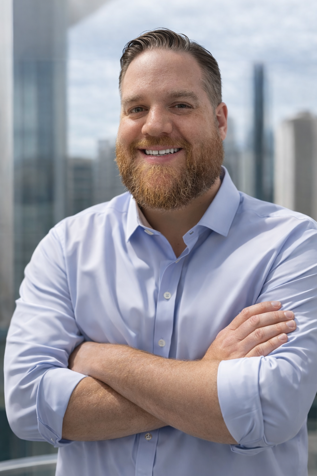 A smiling man with a beard and short, styled hair, wearing a light blue collared shirt with arms crossed, standing in front of a window with a cityscape background.