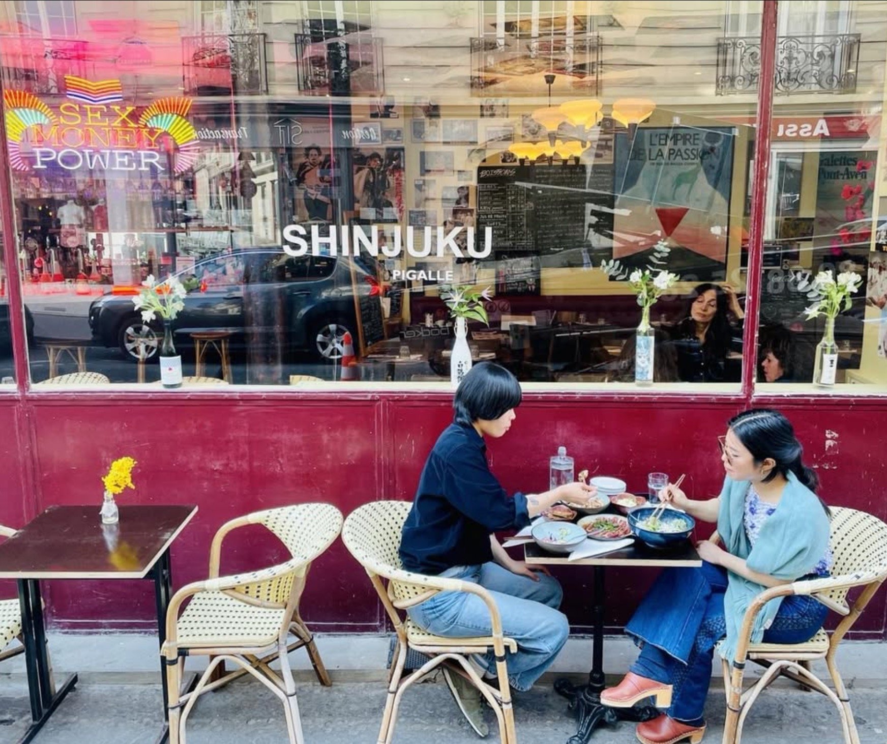 Deux femmes mangent à une table extérieure d'un restaurant à Shinjuku, Tokyo, avec une façade rouge et des fleurs dans des vases. La devanture du restaurant affiche le nom 'SHINJUKU PIGALLE'.