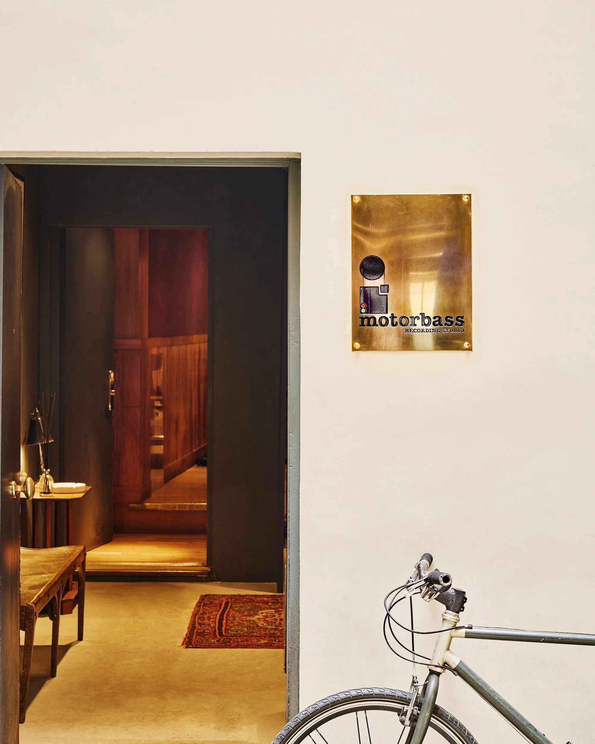 Intérieur d'un studio d'enregistrement nommé Motorbass, porte ouverte, affichage en métal avec le logo du studio. Partiellement visible, un vélo est à l'extérieur.