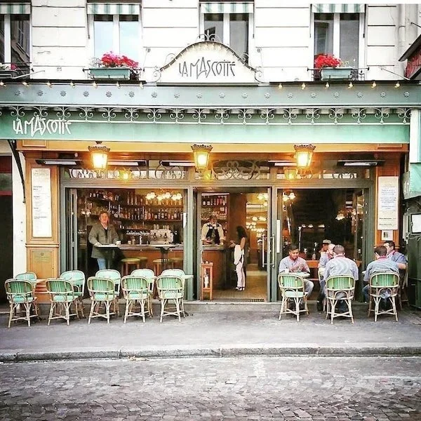 Façade d'un caféou restaurant appelé La Mascotte, avec des clients assis aux tables devant, ouverte sur une rue pavée, avec de la végétation et des lampes éclairant l'entrée.