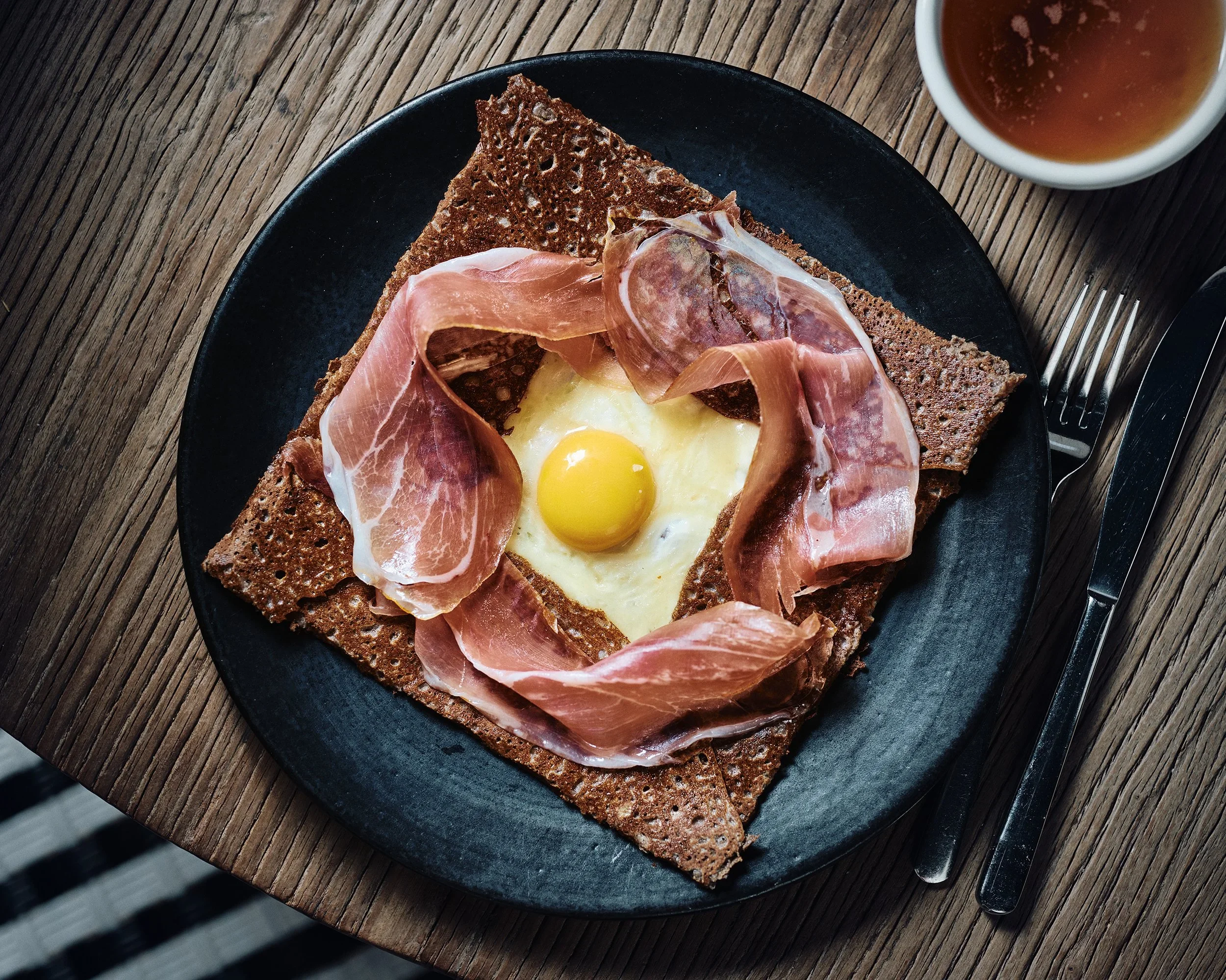 Assiette avec un œuf au plat entouré de fines tranches de jambon sur une tranche de pain noir, accompagnée d'une tasse de thé ou café, sur une table en bois.