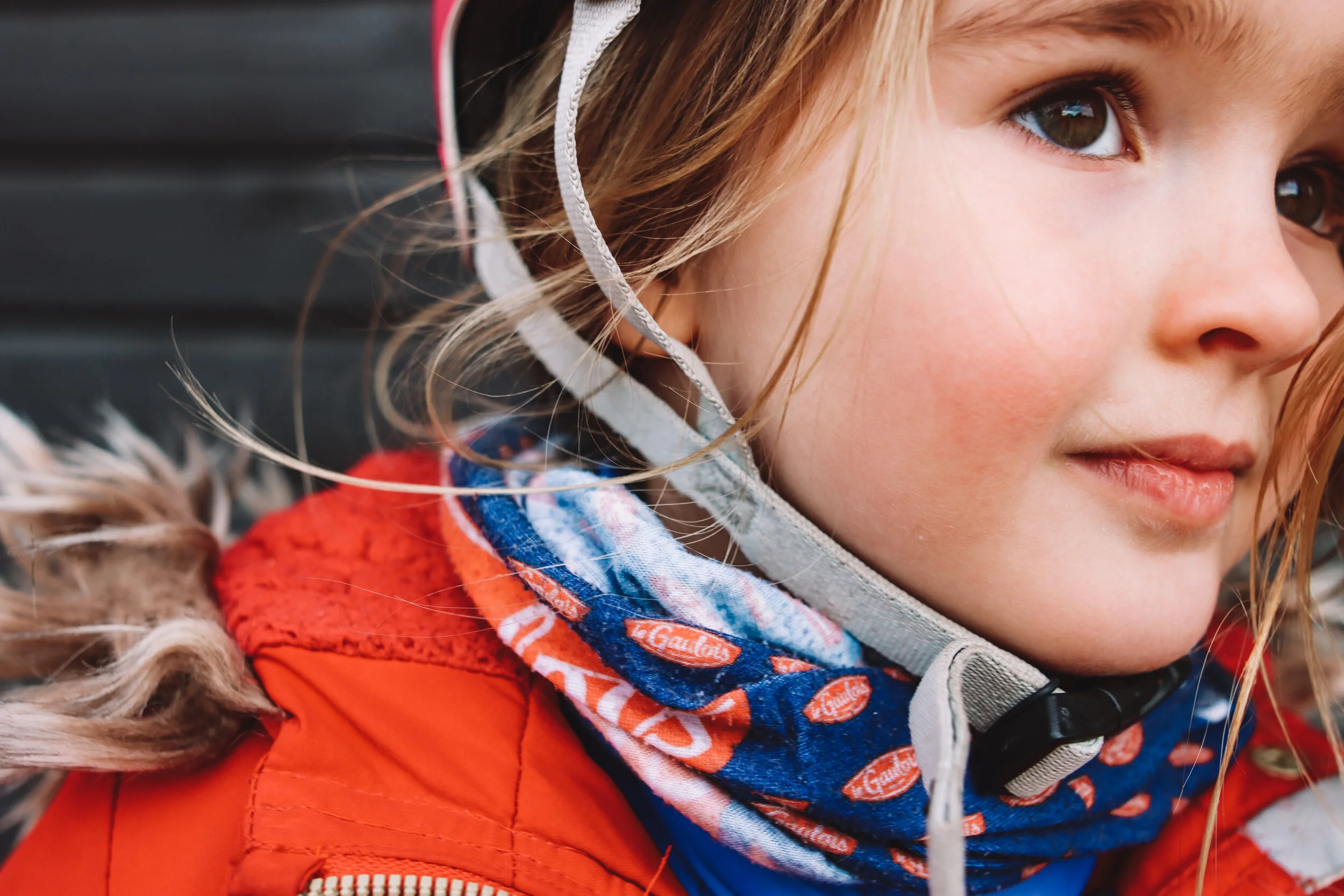 Close-up of a young girl with brown eyes and blonde hair, wearing a helmet, an orange jacket, and a patterned blue scarf, outdoors.