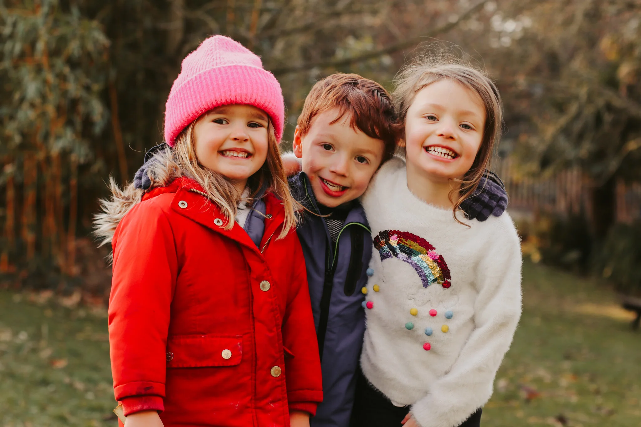 Three children smiling and posing outdoors in autumn, dressed warmly in jackets and sweaters.
