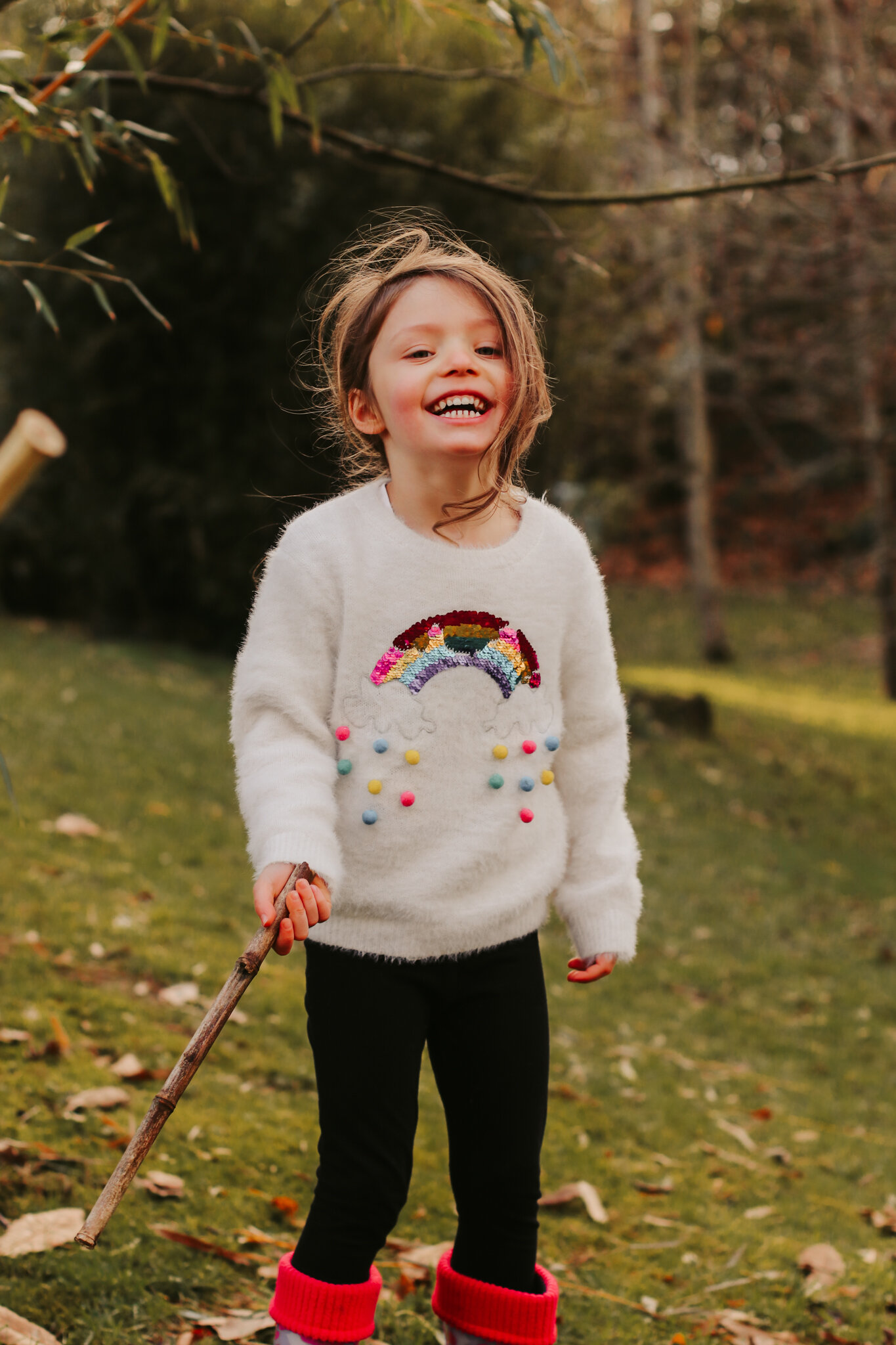 A smiling young girl with light brown hair, wearing a white fluffy sweater with a colorful rainbow and pom-pom design, black pants, and red socks. She is holding a stick and standing outdoors on grass surrounded by trees.
