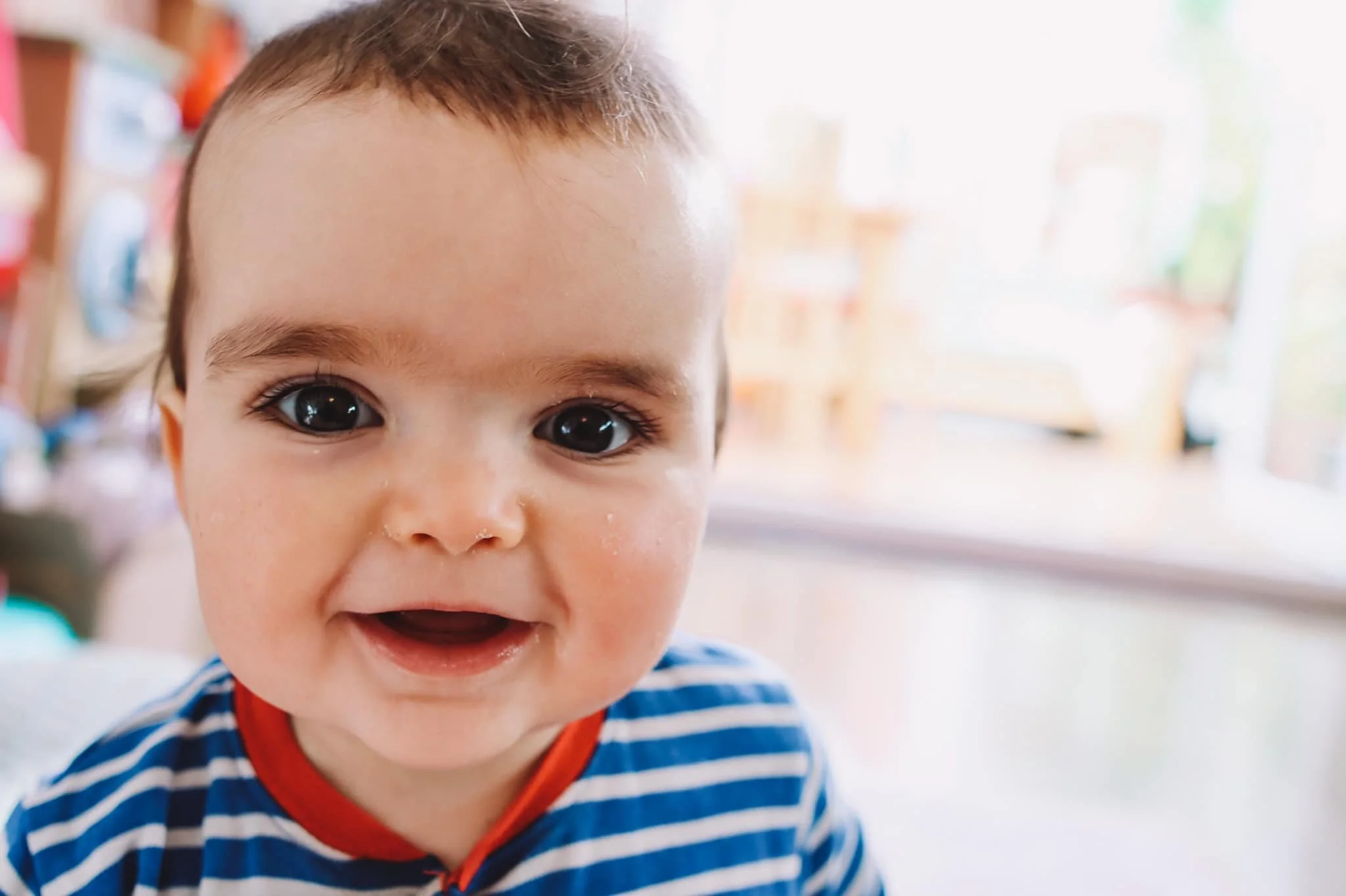Close-up of a smiling toddler with big eyes, wearing a blue and white striped shirt with a red trim, indoors with a blurred background