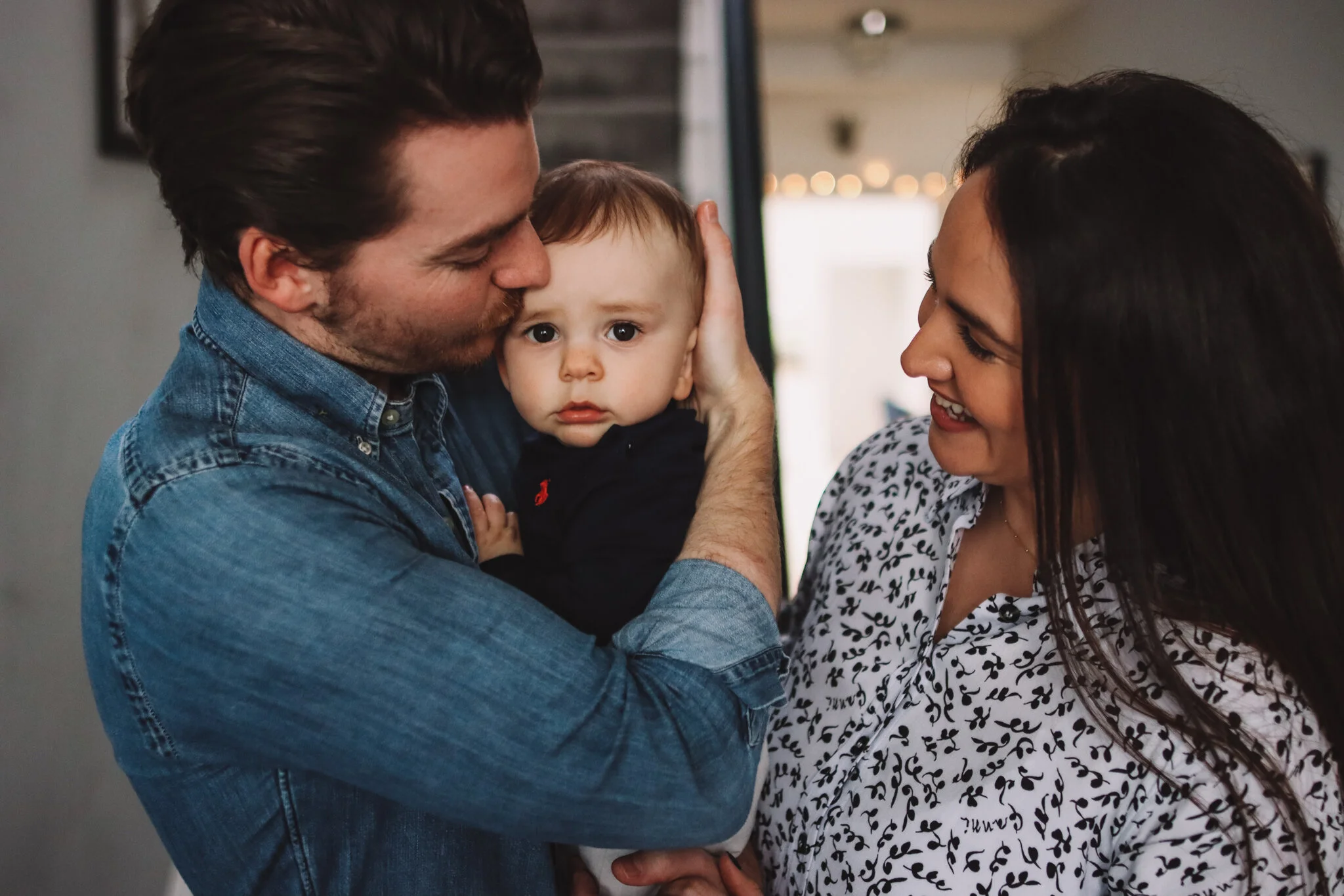 A family scene with a man, woman, and a young child. The man is kissing the child's forehead, the woman is smiling at the child, and the child looks surprised or curious. They are indoors, near a window and casual surroundings.