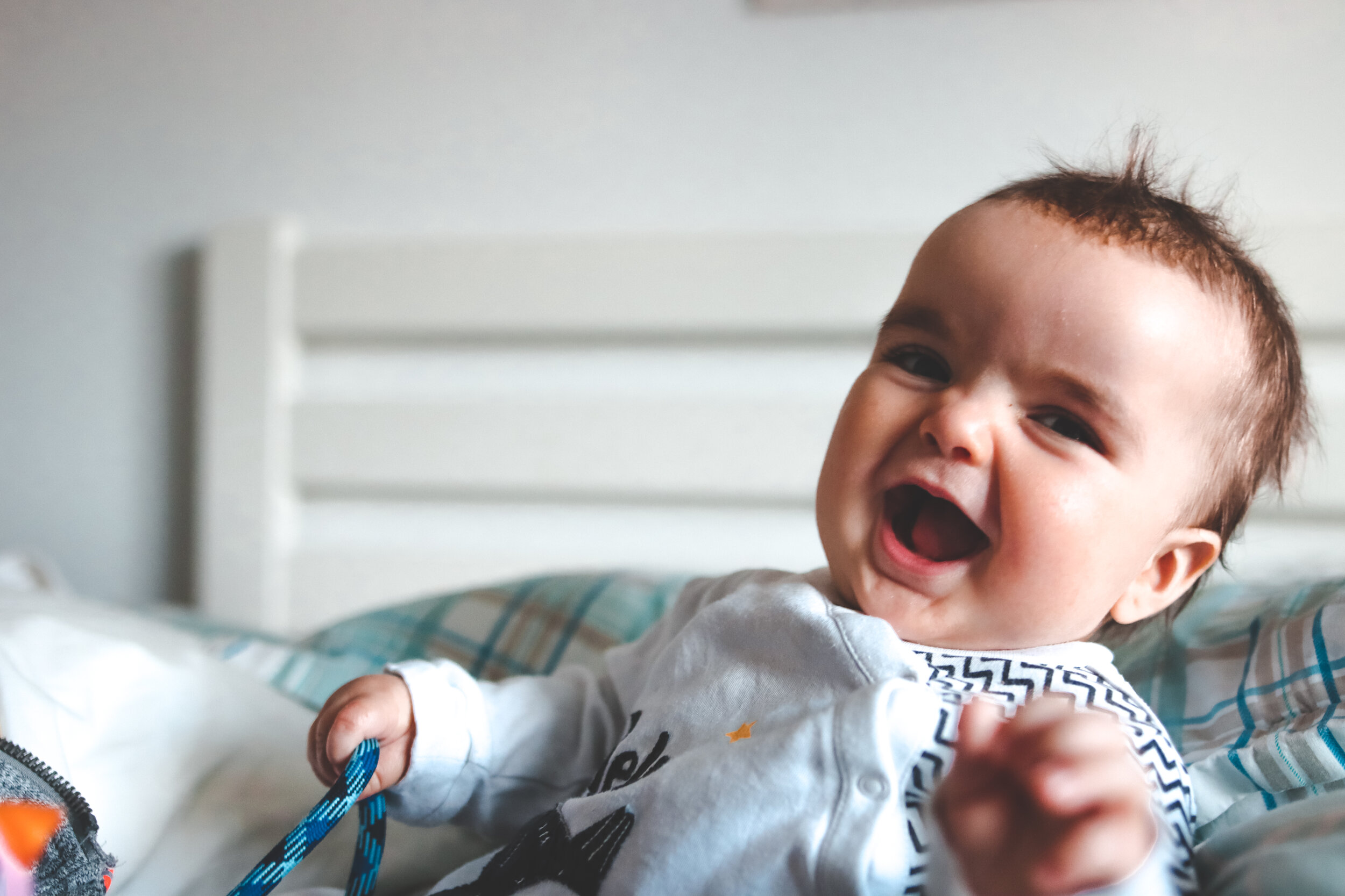 A happy baby with a big smile, lying on a bed, holding a blue and black striped object in one hand.