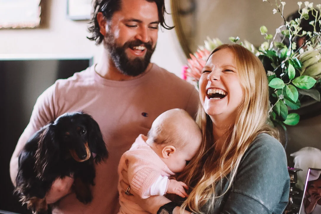 A happy family with a man, woman, a baby, and a dog, all smiling and laughing indoors.