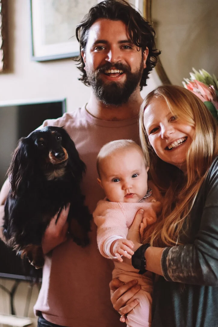 A happy family of three people and a dog, smiling and posing together indoors. The man has dark hair and a beard, the woman has long blonde hair, and they are holding a baby girl with light hair. The dog is black with brown markings.