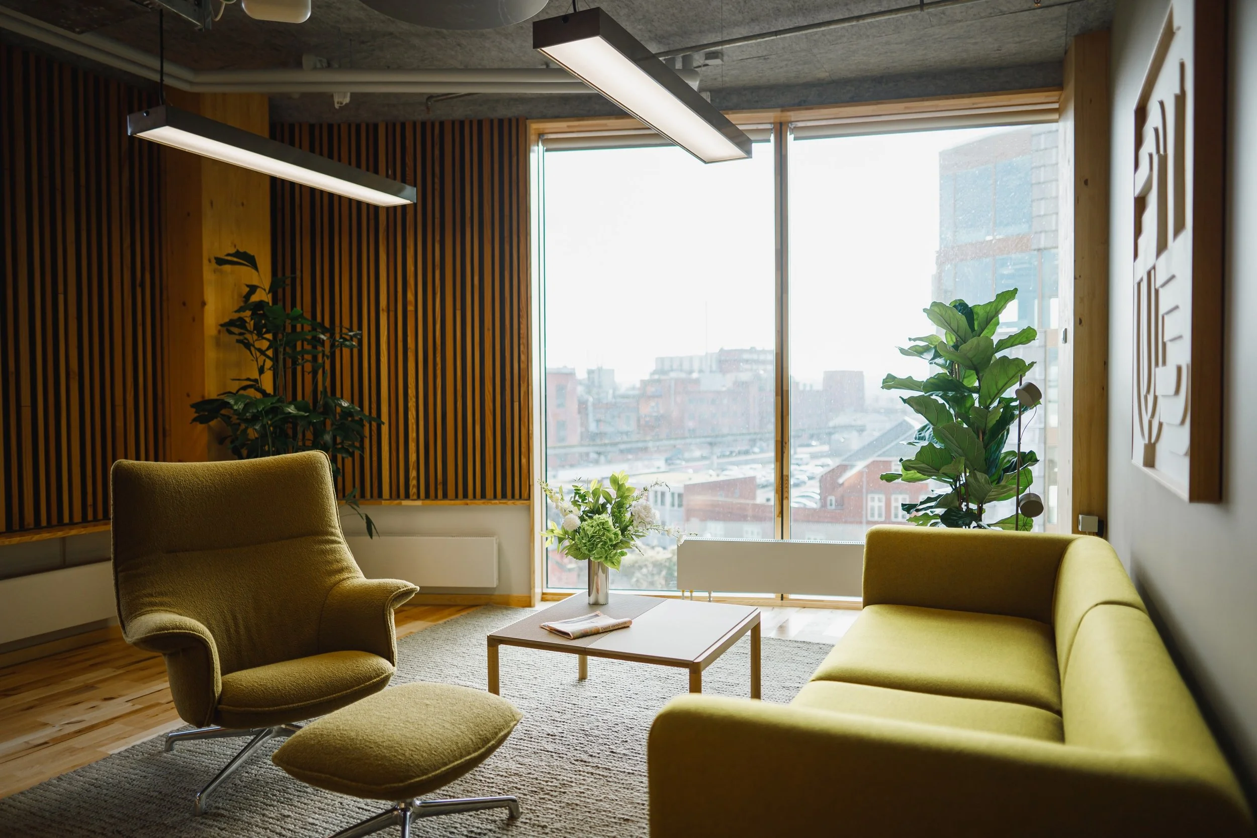 Indendørs moderne kontorlokale med store vinduer, grønne planter og loungeområder med lysegrønne sofaer.
