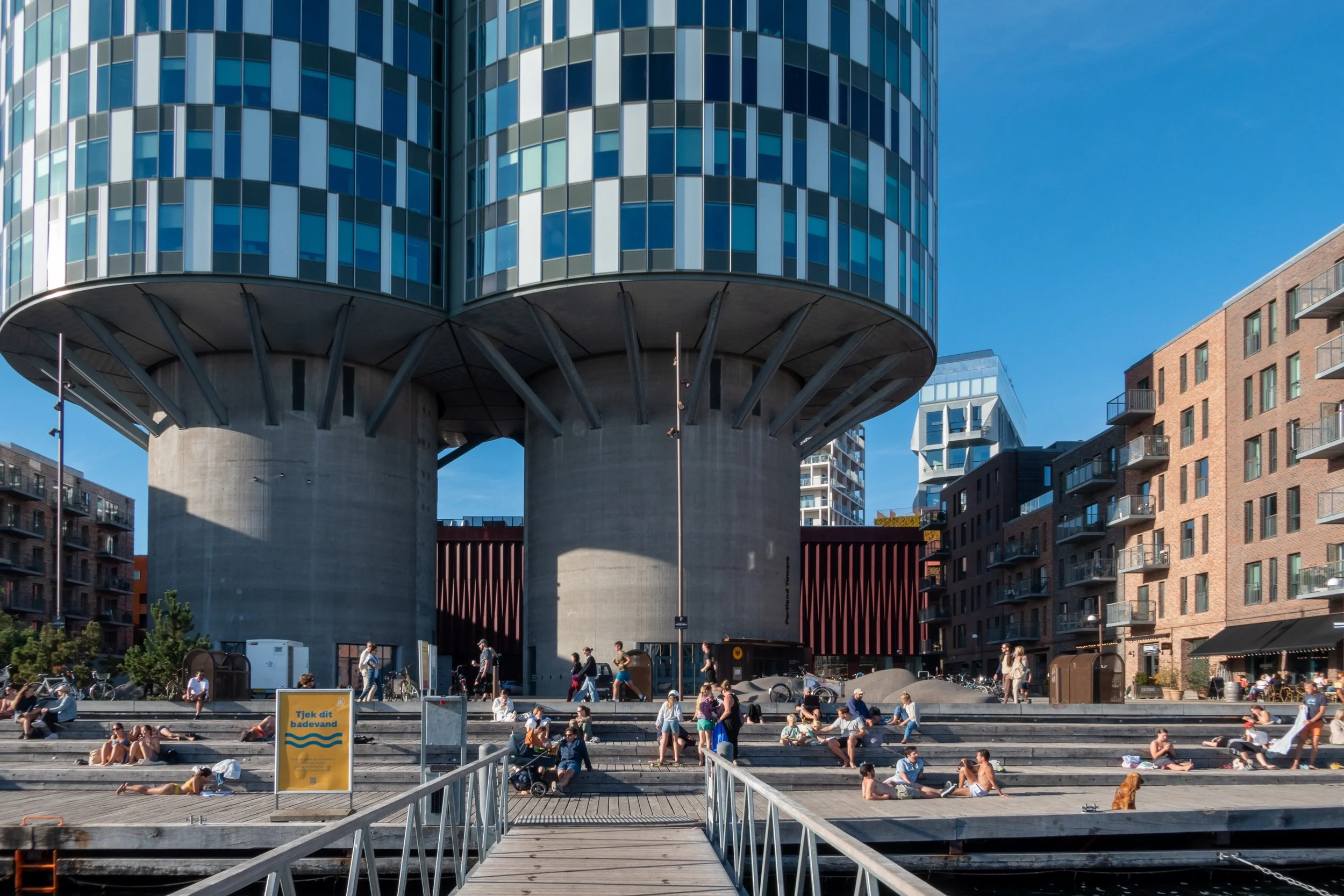 Moderne bygning med en rund, hævet del, der er støttet af store betonpiller. Beboelsesbygninger med balkoner er synlige til højre. Mange mennesker nyder vejret på trappesperspektivet foran bygningen, nogle sidder og nogle går rundt. En skilt, der opfordrer til at tjekke badetøj, er synligt.