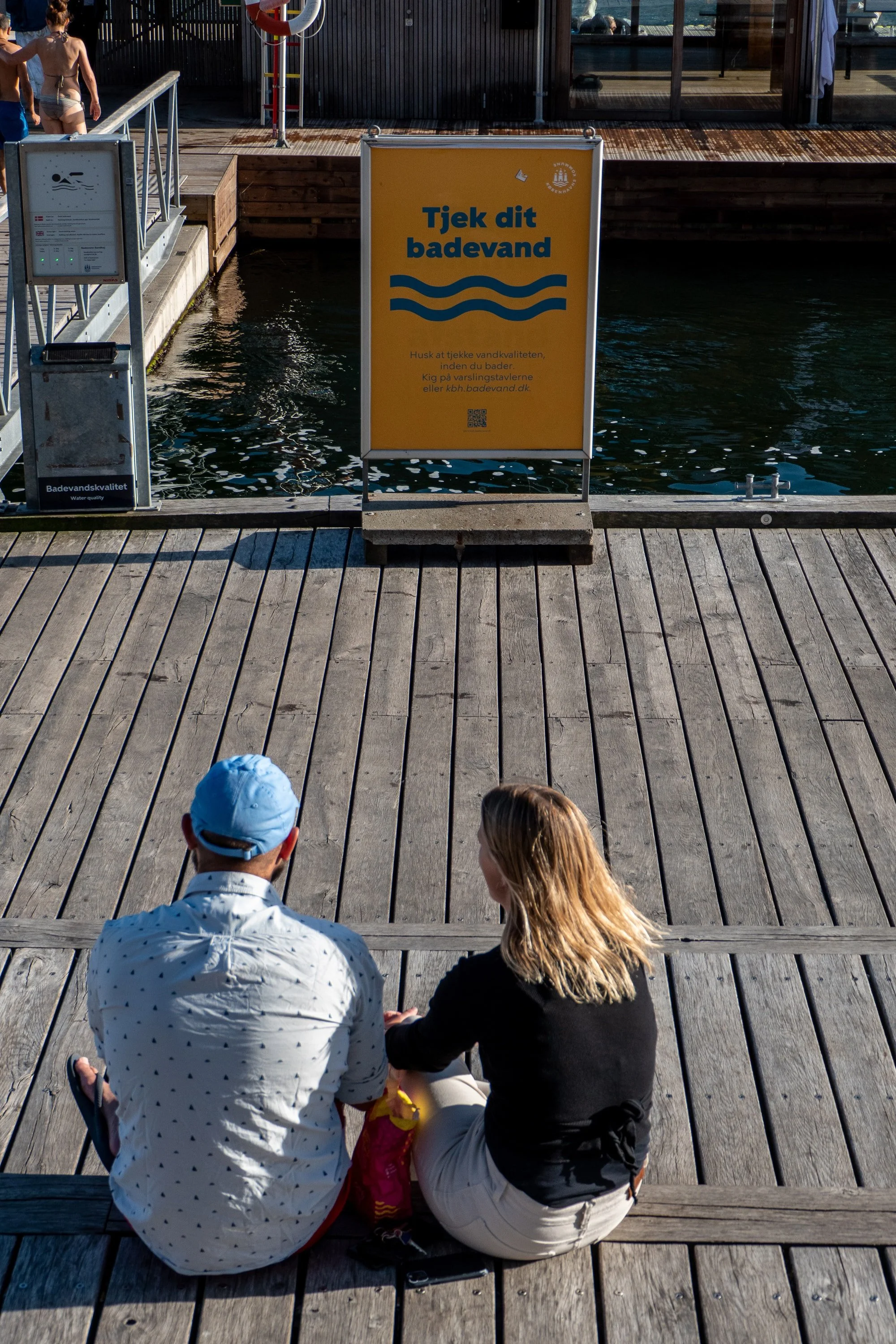 To personer sidder på en trædæk foran en vandkant, der ser ud til at være ved en pool eller et vandreservoir. En reklameskilt med teksten 'Tjek dit badevand' er placeret foran dem, og to børn står i vandet i baggrunden.