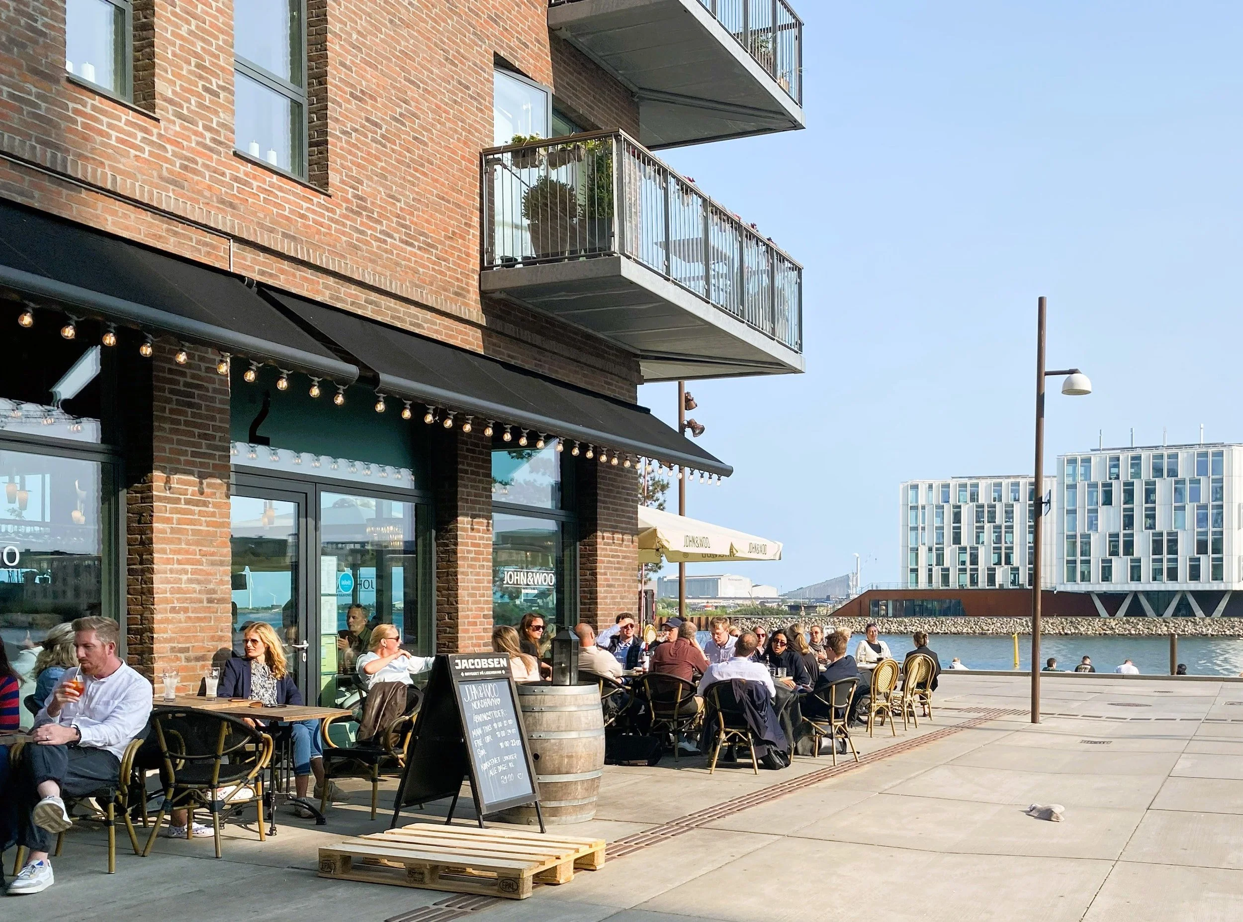 Udendørs restaurant med mange gæster, der spiser og drikker ved borde foran en bygning med store vinduer og altaner. Der er en sort tavle med menukort og en stor fad med reklame til en restaurant. Ibaggrunden ses en mole og en moderne bygning med mange vinduer, samt en datalampe på en stolpe mod den blå himmel.