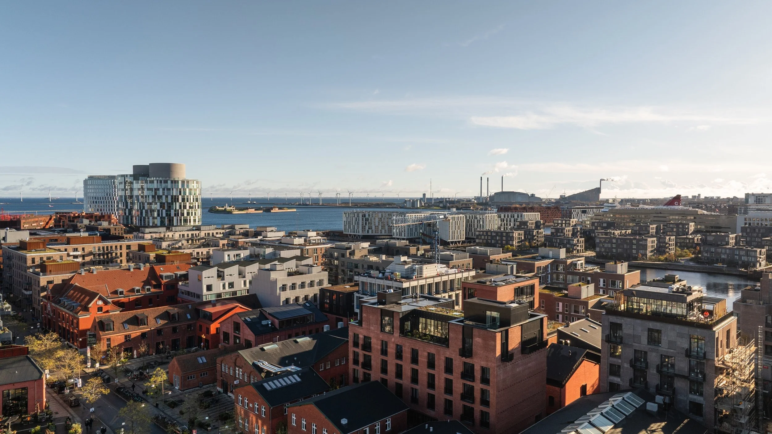 Udsigt over en by midt i byen med moderne bygninger, en havn med skibe, vindmøller i baggrunden og blå himmel.