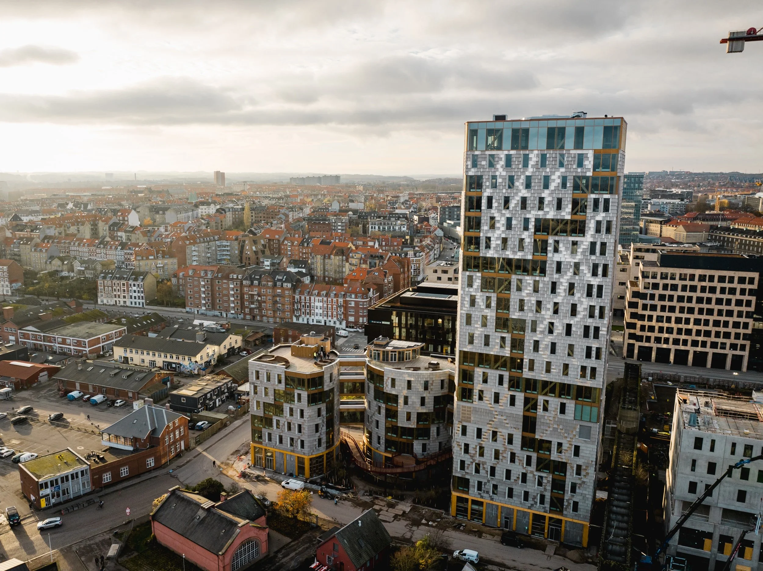Billedet viser en by med forskellige bygninger, herunder højhuse og lavere bygninger. Der er en moderne højopført bygning med et mærkeligt mønster og store vinduer i forgrunden. Baggrunden viser en palette af gamle og nye bygninger under en overskyet himmel.