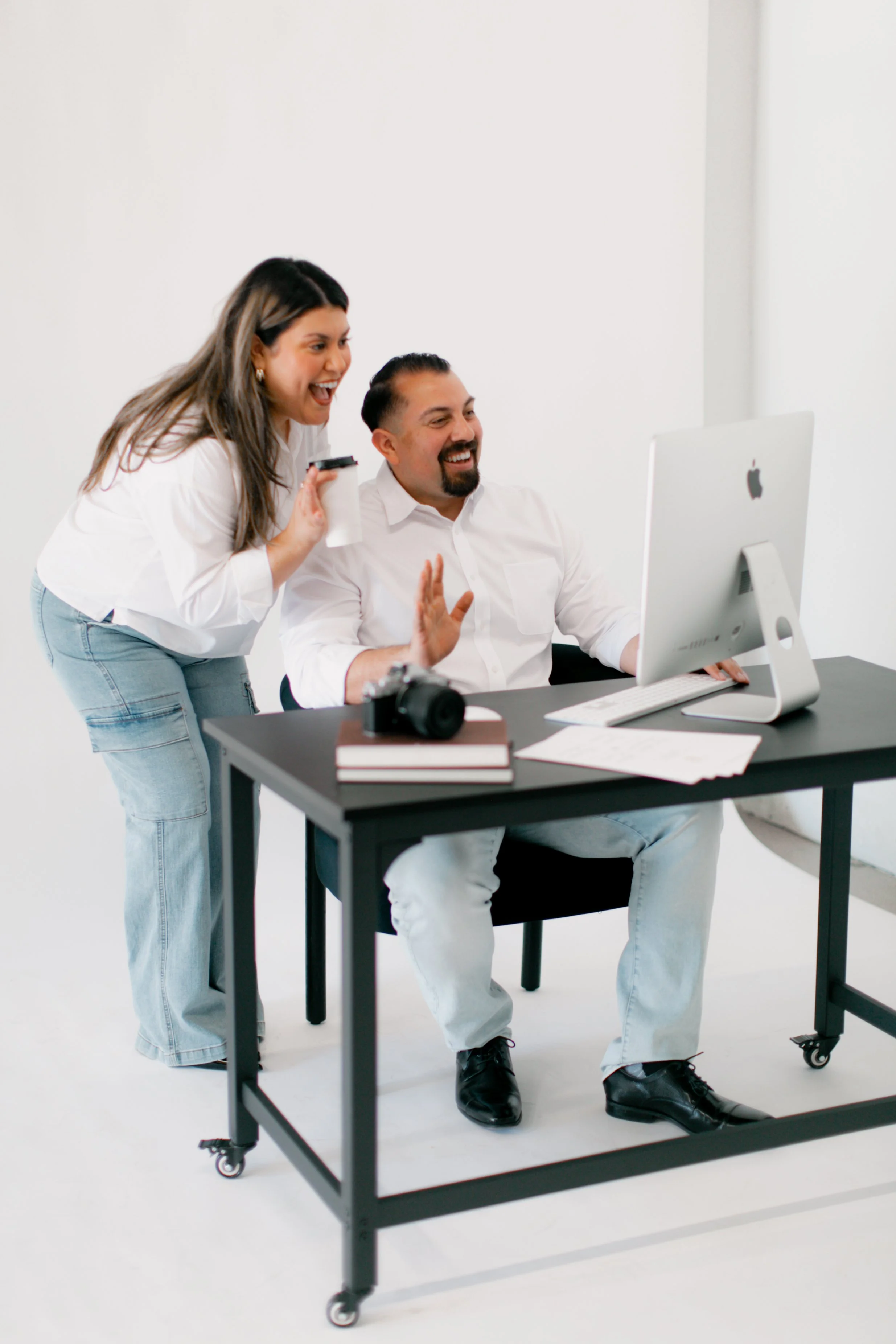 Two coworkers, a woman and a man, are laughing and smiling while looking at a computer screen in an office setting. The woman holds a coffee cup.