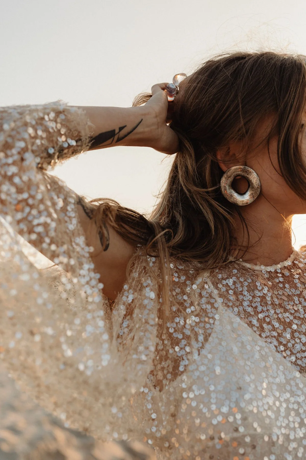 A woman with brown hair, large hoop earrings, and a sequin dress, touching her hair with her left hand. Her face is turned away from the camera. The background is neutral and the lighting is soft.