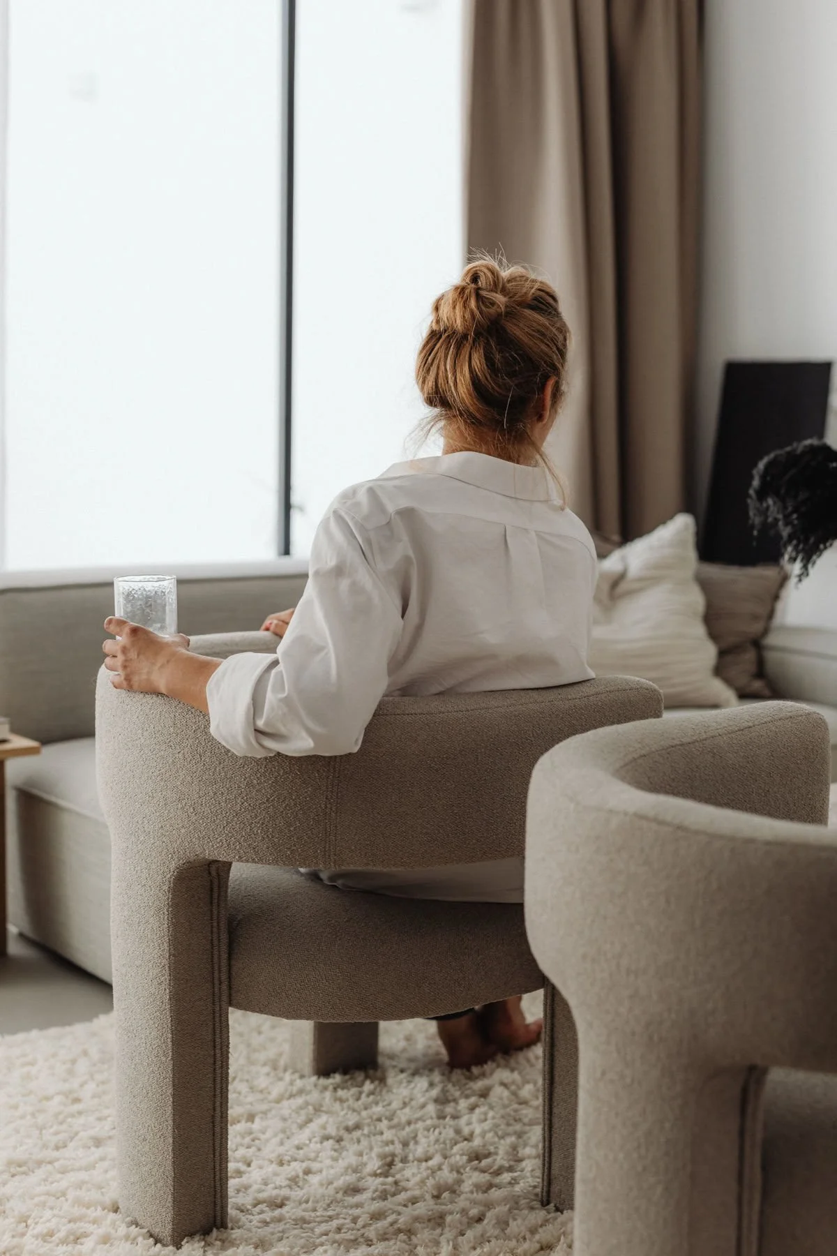 A woman sitting on a beige upholstered armchair in a modern living room, holding a glass of water in her right hand, with her back to the camera, gazing towards a large window with brown curtains.