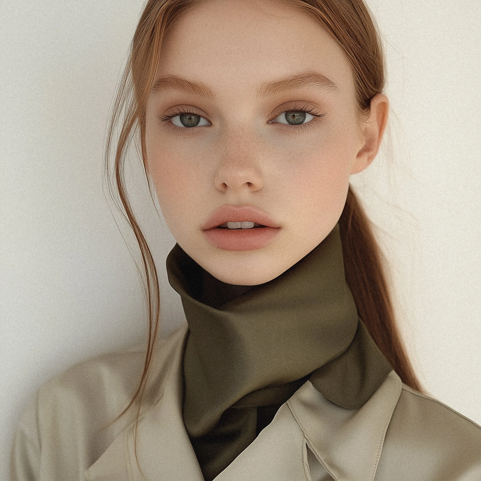 Portrait of a young woman with red hair, blue eyes, and natural makeup, wearing a beige trench coat and an olive green scarf against a neutral background.