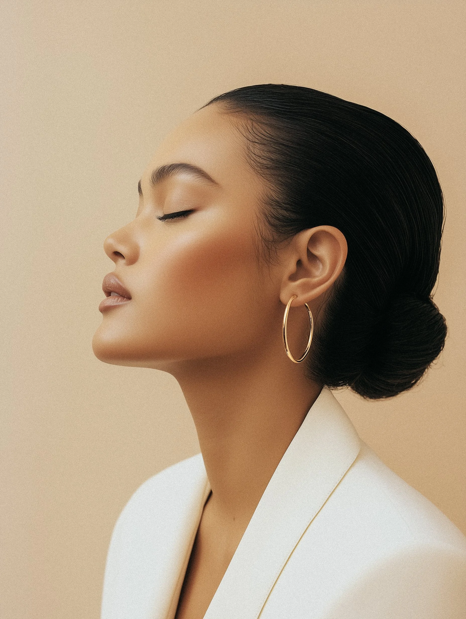 Side profile of a woman with dark hair styled in a low bun, wearing gold hoop earrings and a white blazer, with a neutral beige background.