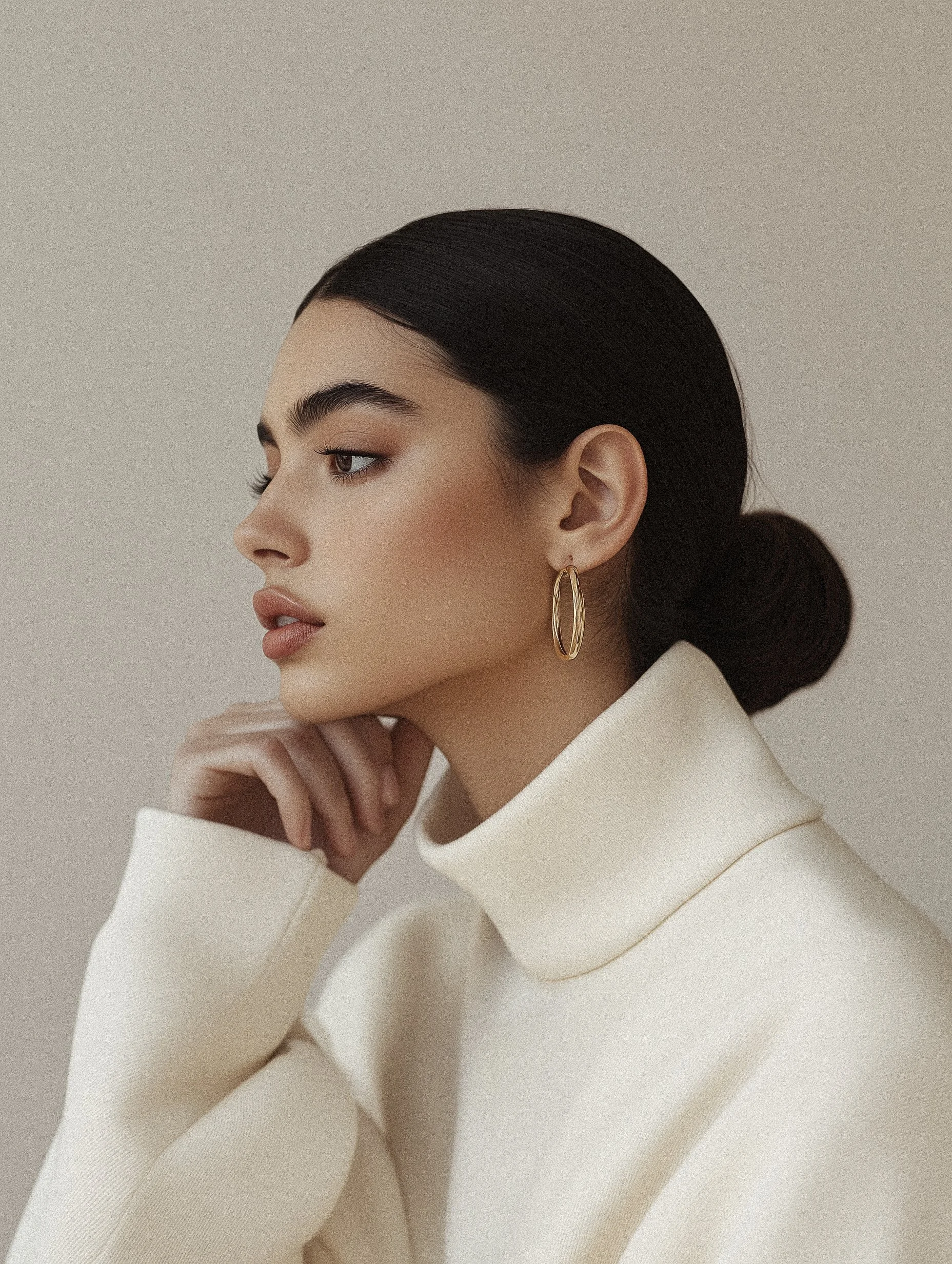 A woman with dark hair in a bun, wearing a cream turtleneck sweater and gold hoop earrings, is shown in profile against a neutral background.