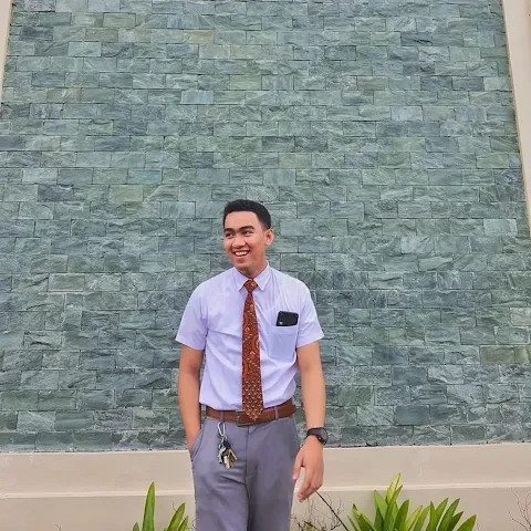 Young man in business casual attire smiling outdoors against a textured green wall.