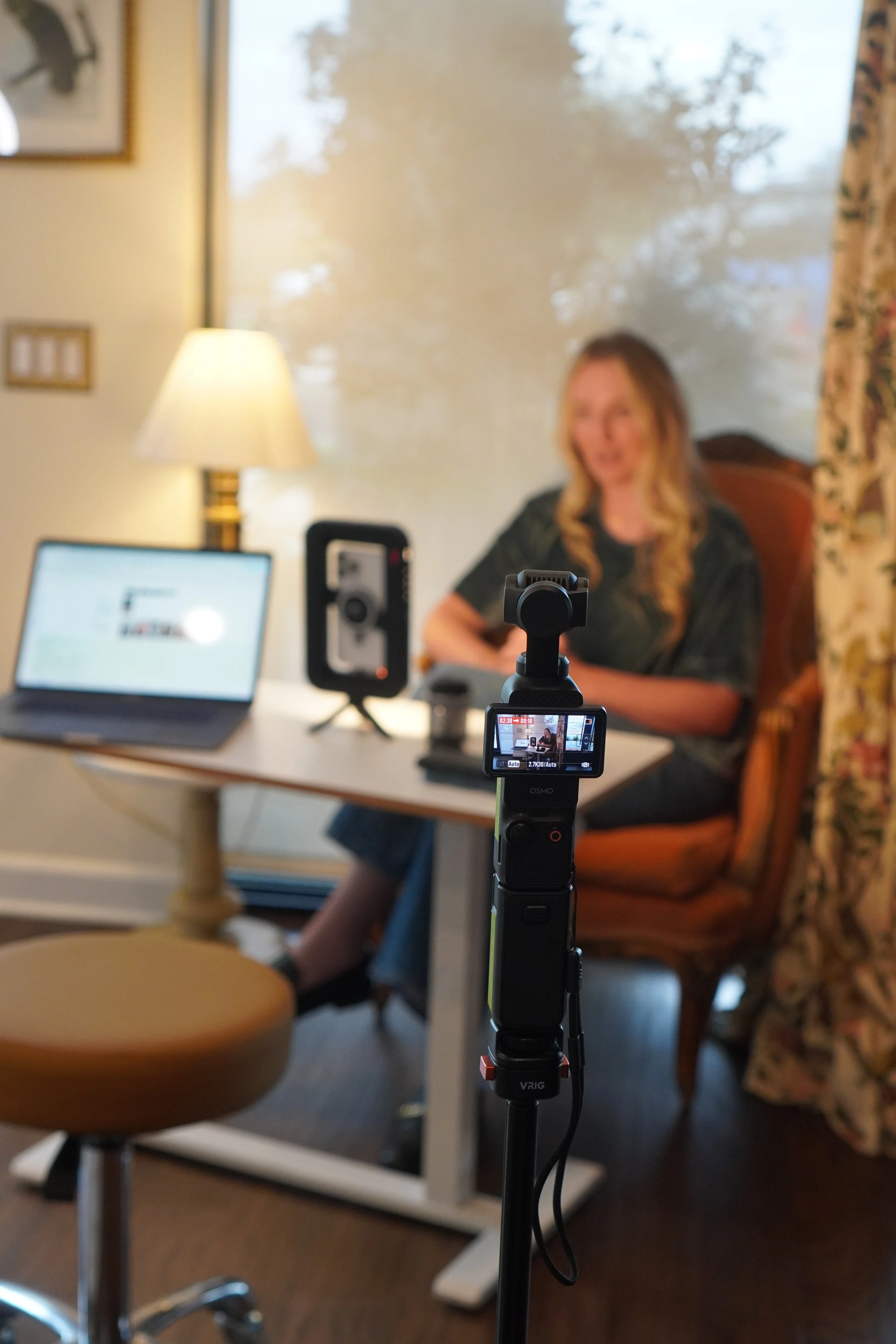 A woman sitting on an orange armchair in a room, with a lamp, a laptop, and a speaker on a table in front of her. The photo is taken through a camera mounted on a tripod, capturing her in focus.