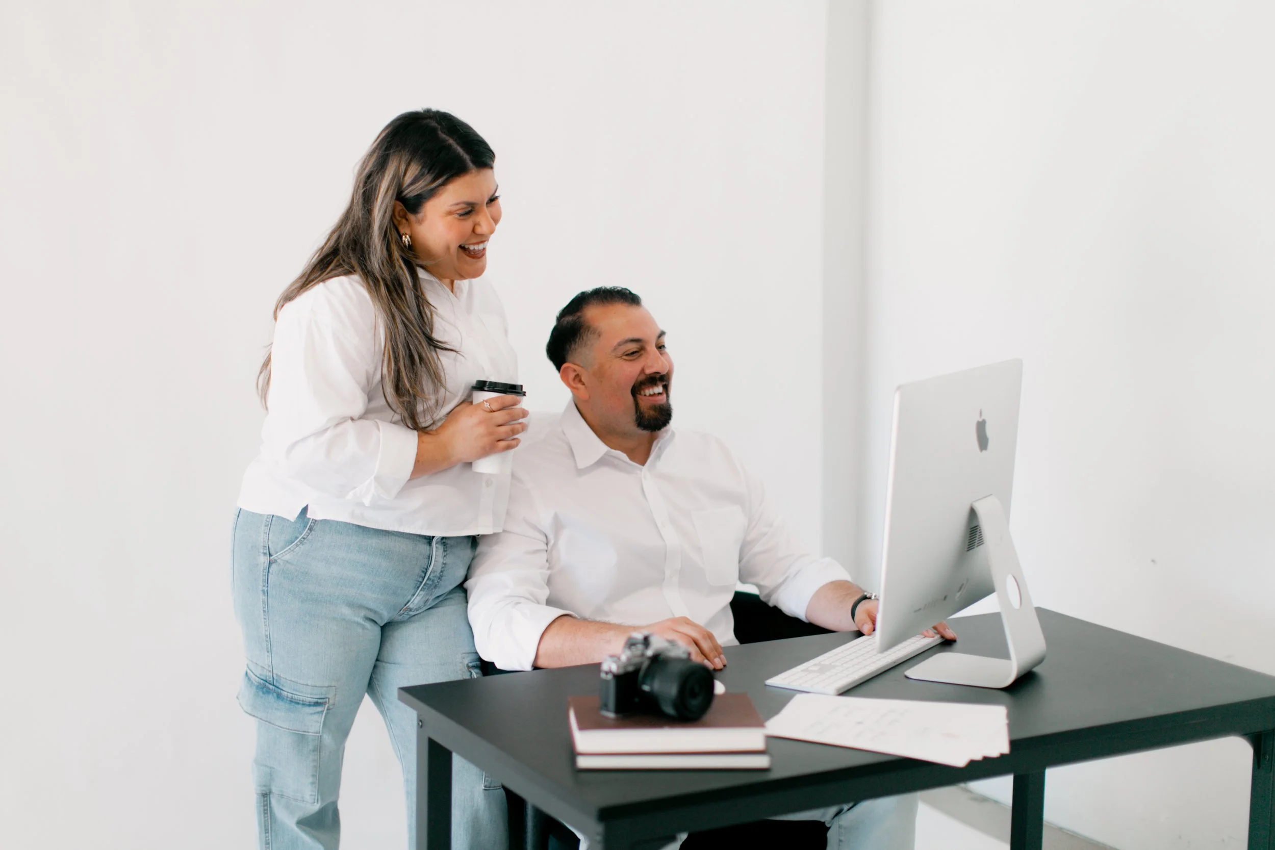 A woman and a man are working together at a desk in an office. The woman is standing and holding a coffee cup, smiling at the man who is sitting at a computer. There is a camera, books, and papers on the desk.