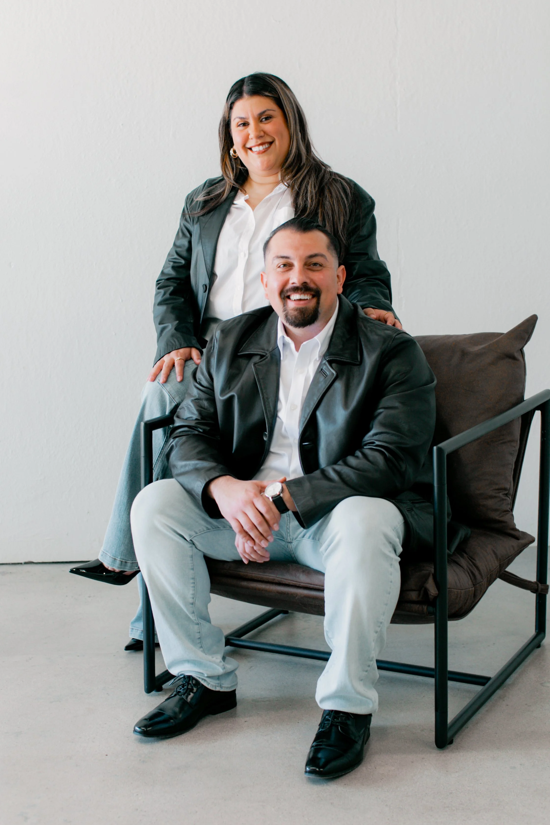 A woman and a man sitting and standing together in a modern room, both smiling and wearing black leather jackets.