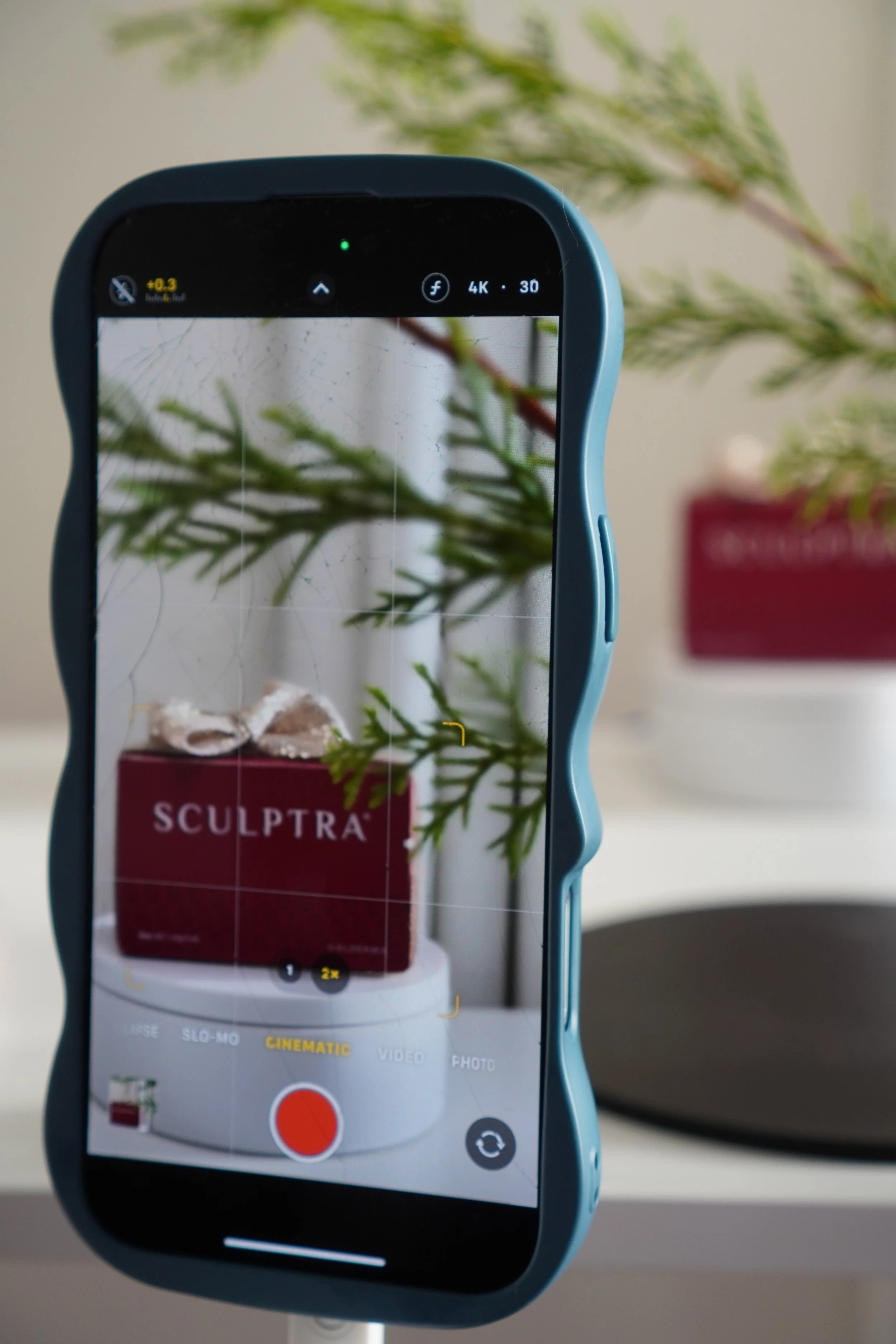 Close-up of a smartphone on a tripod capturing a photo of a decorative arrangement on a round white base with a red box labeled 'SCULPTRA' and seashells on top, with greenery in the background.