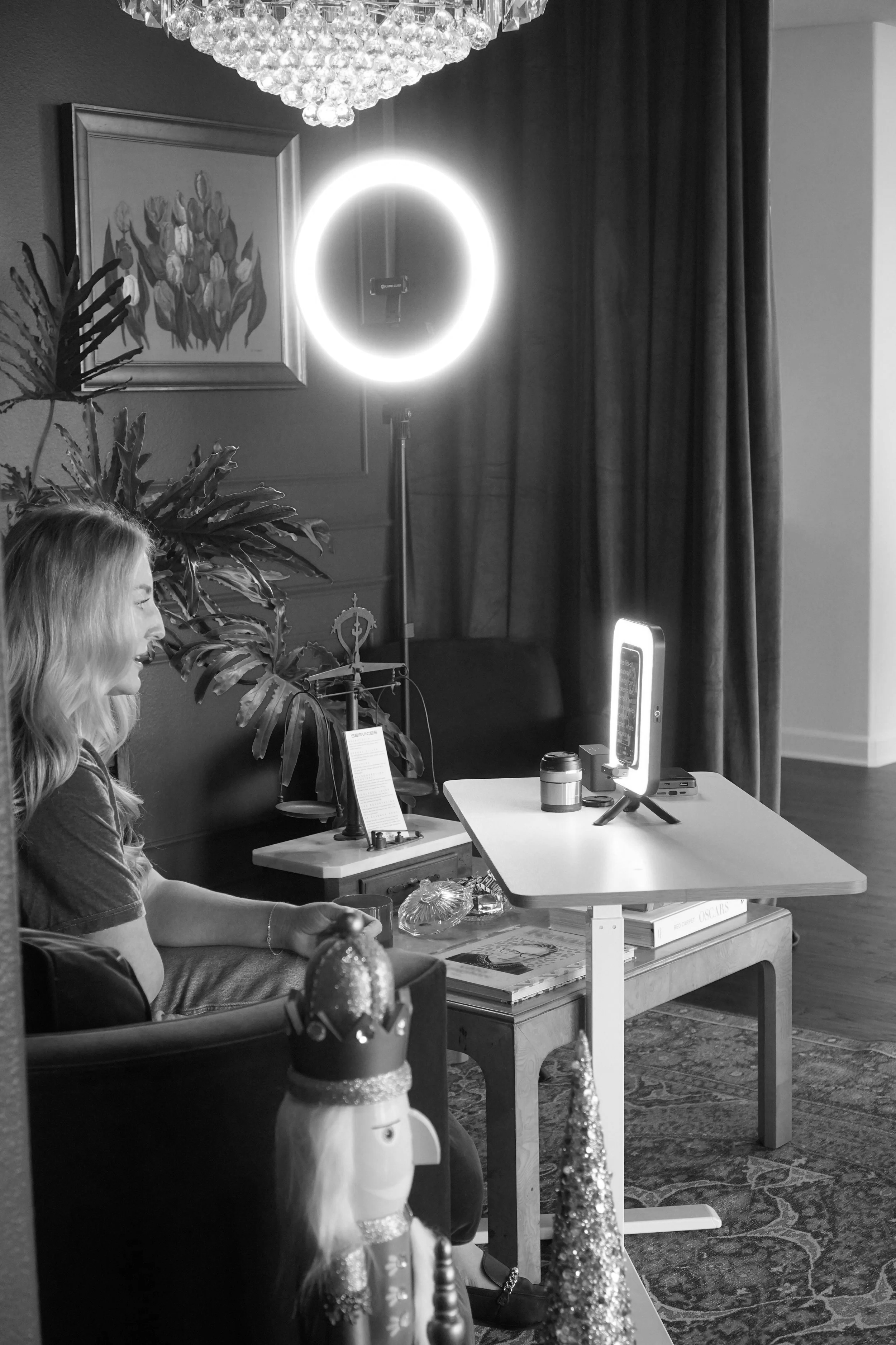 A woman sitting in a living room with a laptop, ring light, camera, and decoration, during a video call or recording session; black and white photo.