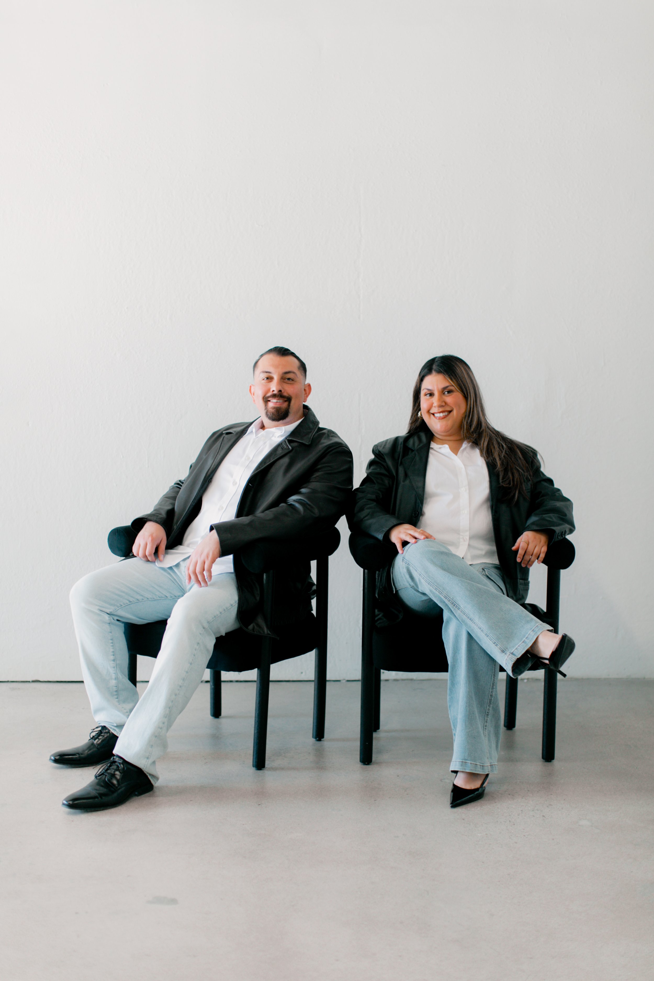 A man and a woman sitting on black chairs against a plain white wall, smiling, wearing casual business attire with black jackets and white shirts.