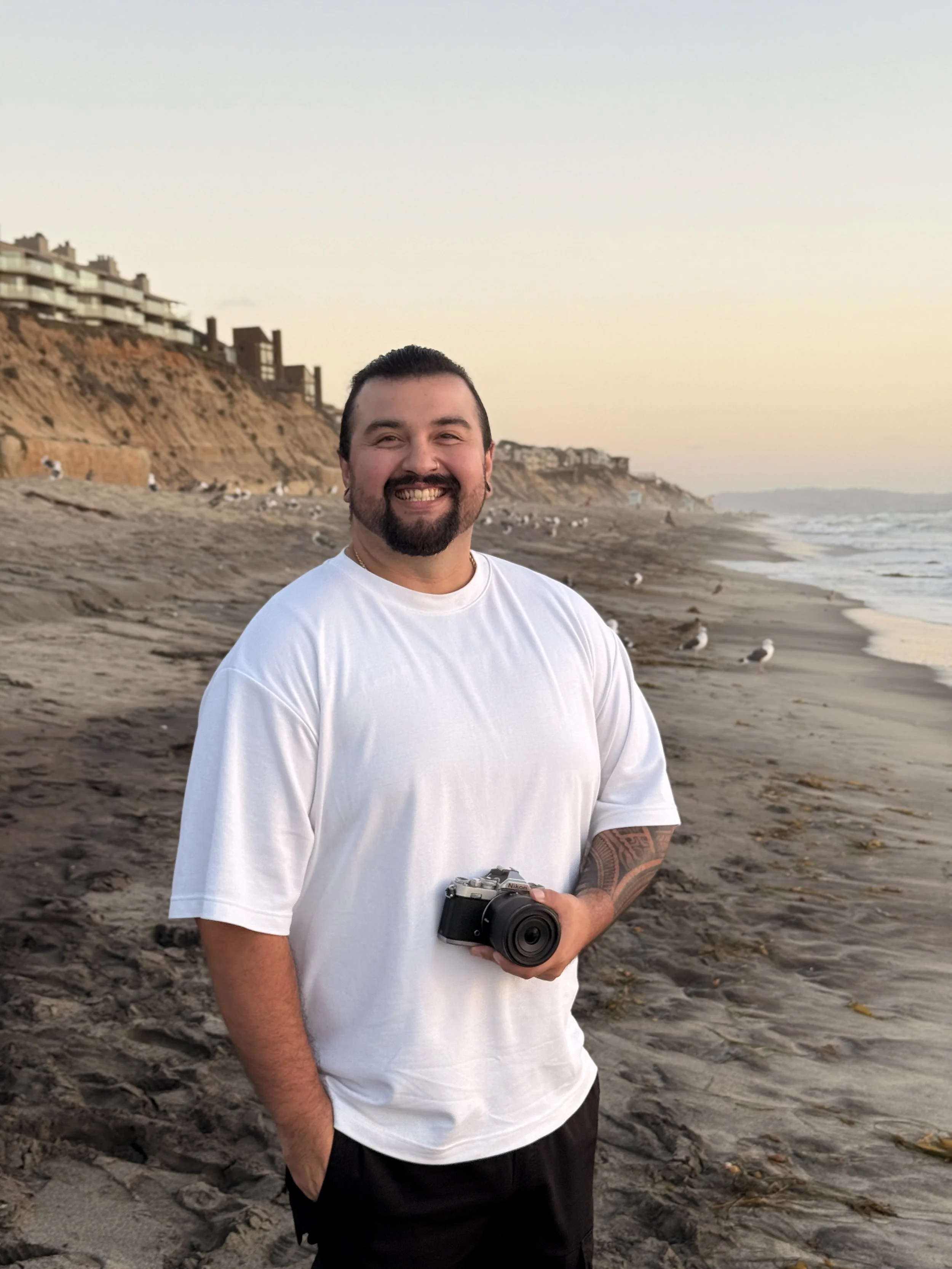 A man with a camera standing on a sandy beach with seagulls, cliffs, and buildings in the background during sunset.