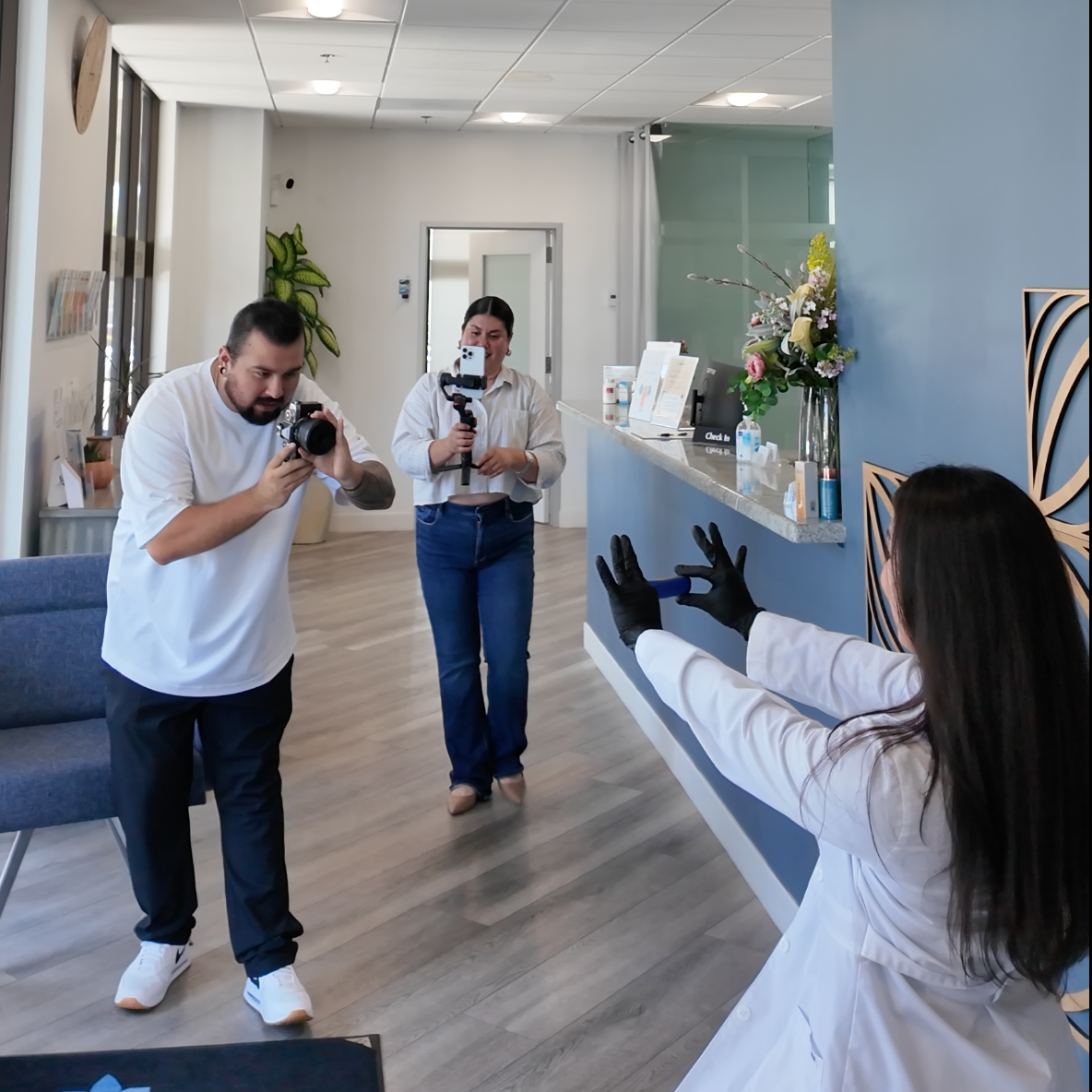 A woman wearing a white lab coat and black gloves is gesturing as she is being filmed by a man and woman filming her with a camera and a smartphone in a modern interior space.