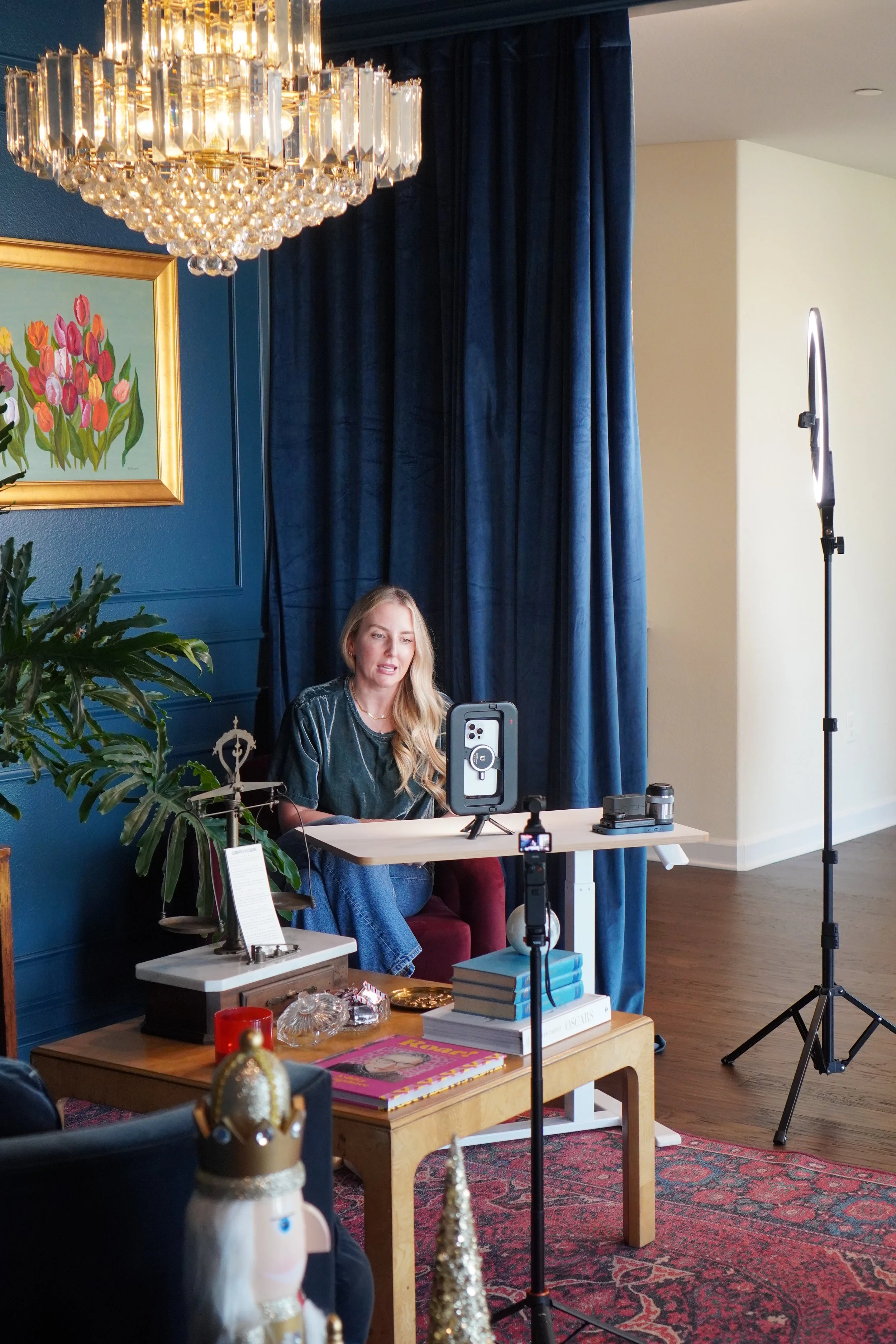 A woman sitting at a small table in front of a blue wall, with a blue curtain to her right, surrounded by video recording equipment and decorative items.