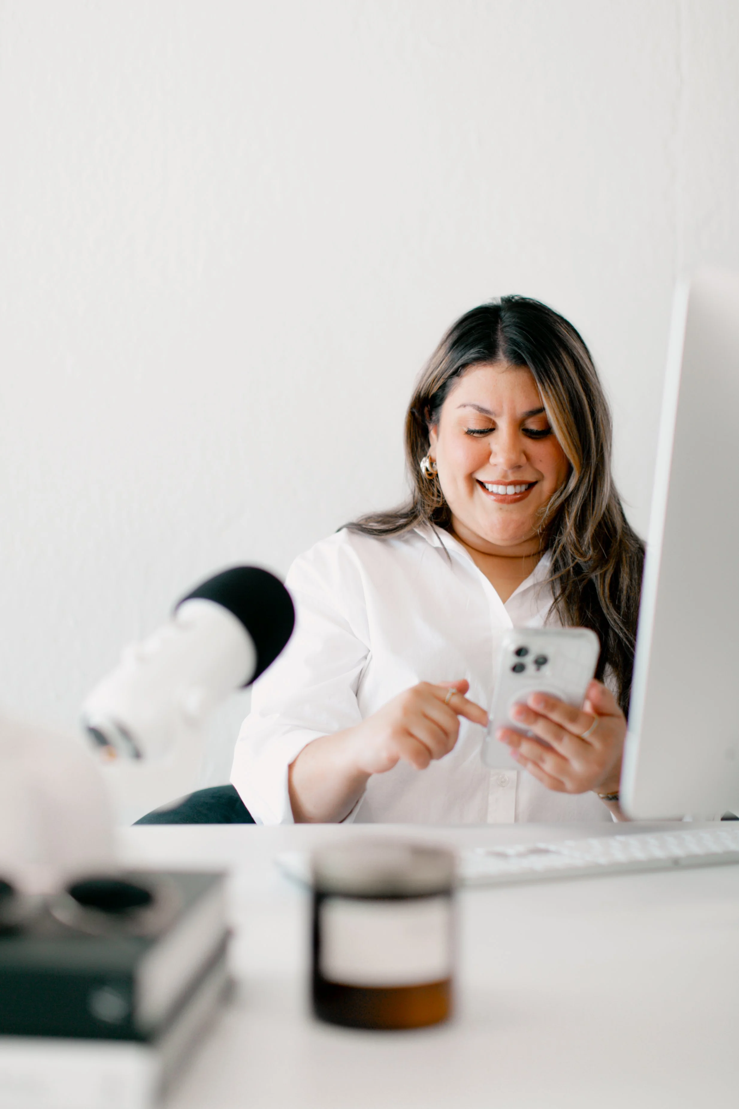 A woman using her smartphone at a desk with a microphone, computer, and a jar, smiling and looking at her phone.
