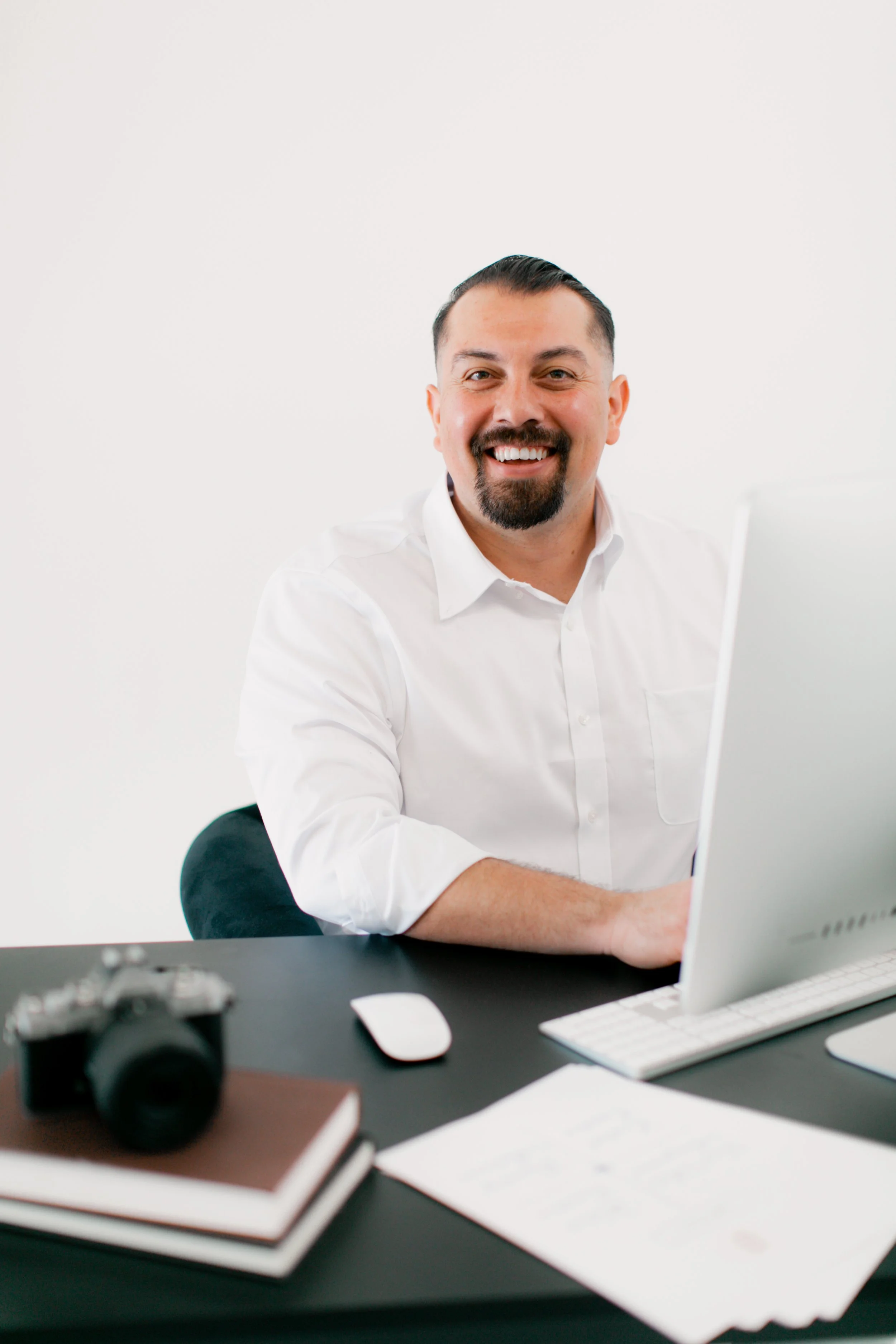 A smiling man sitting at a desk with a computer, camera, notebooks, and papers, dressed in a white shirt.