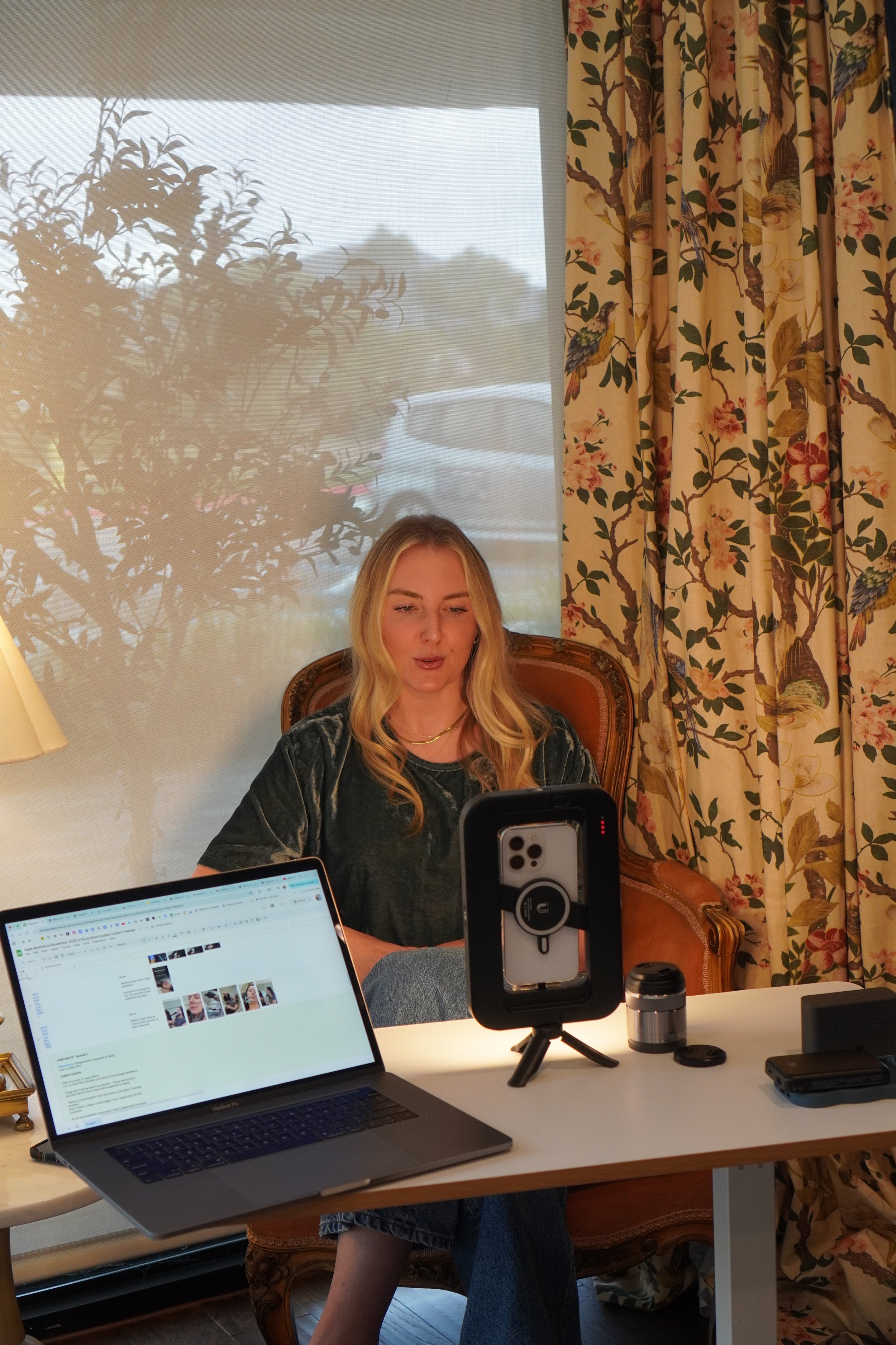 A woman sitting at a desk with a laptop, smartphone on a stand, and various tech devices, in a room with floral curtains and a window with outdoor scenery.